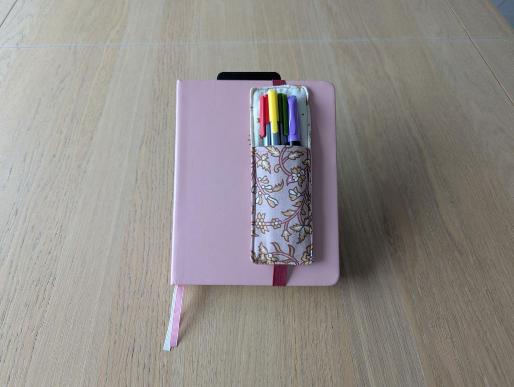 A pink notebook with a pen holder containing colorful pens is placed on a wooden surface.