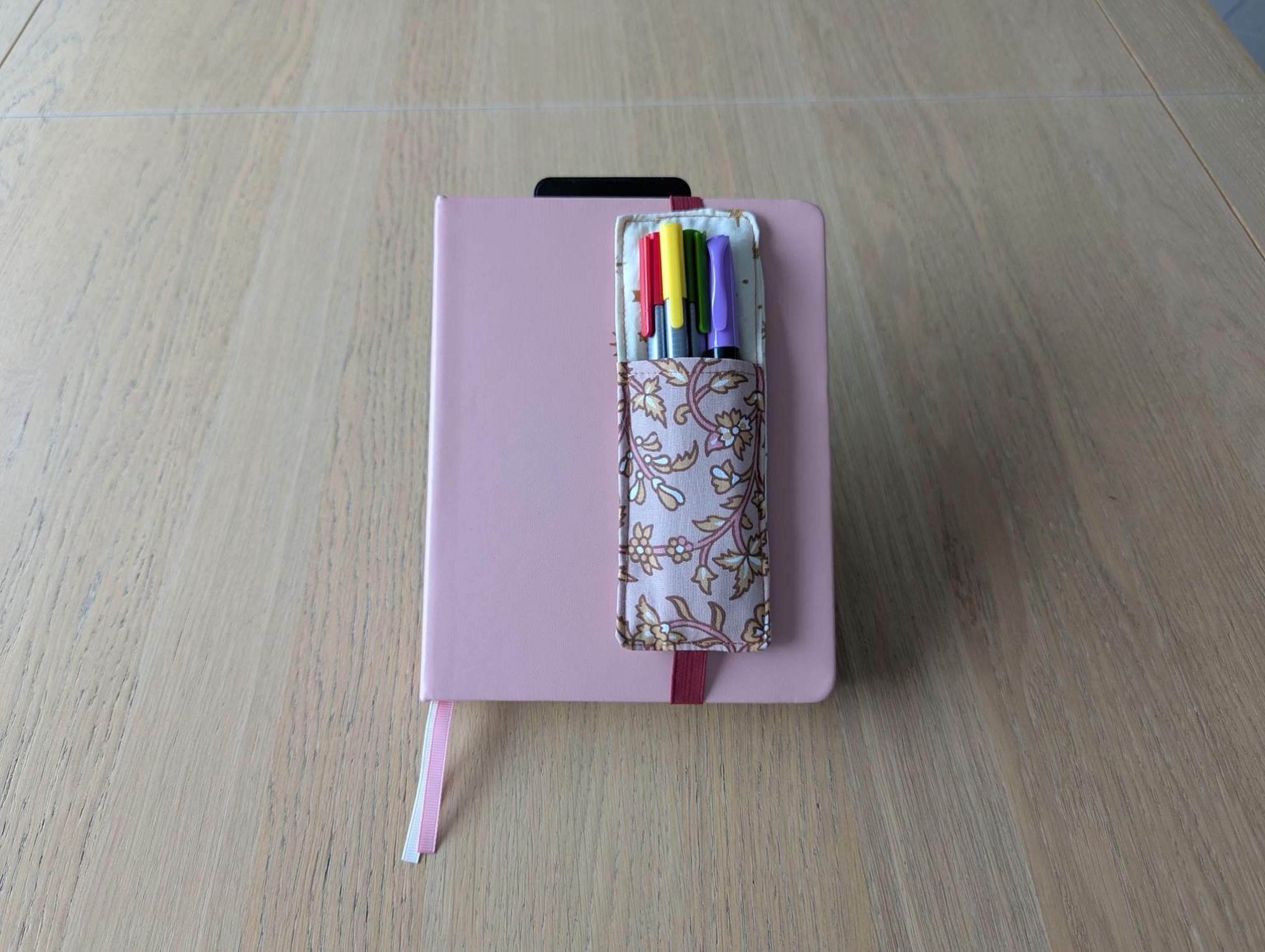 A pink notebook with a pen holder containing colorful pens is placed on a wooden surface.