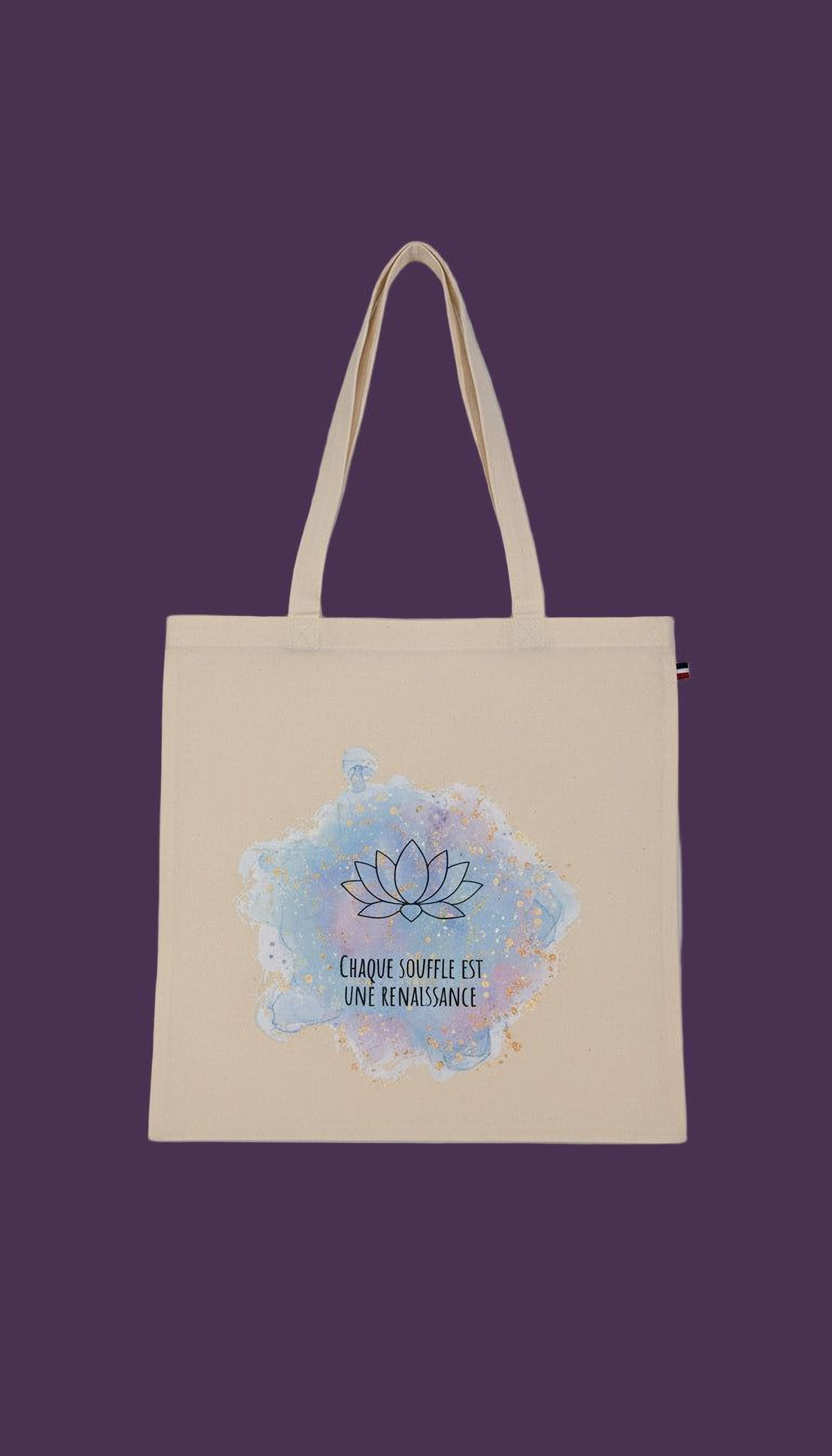 A beige tote bag with a blue and white watercolor design featuring a lotus flower and the text "Chaque souffle est une renaissance" (The lotus is a symbol of renaissance).