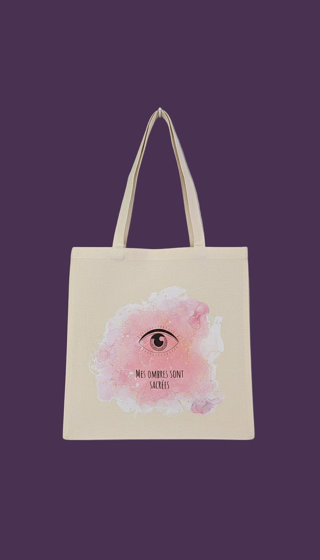 A beige tote bag with a pink eye design and the text "Mes ombre sont sacre" printed on it.