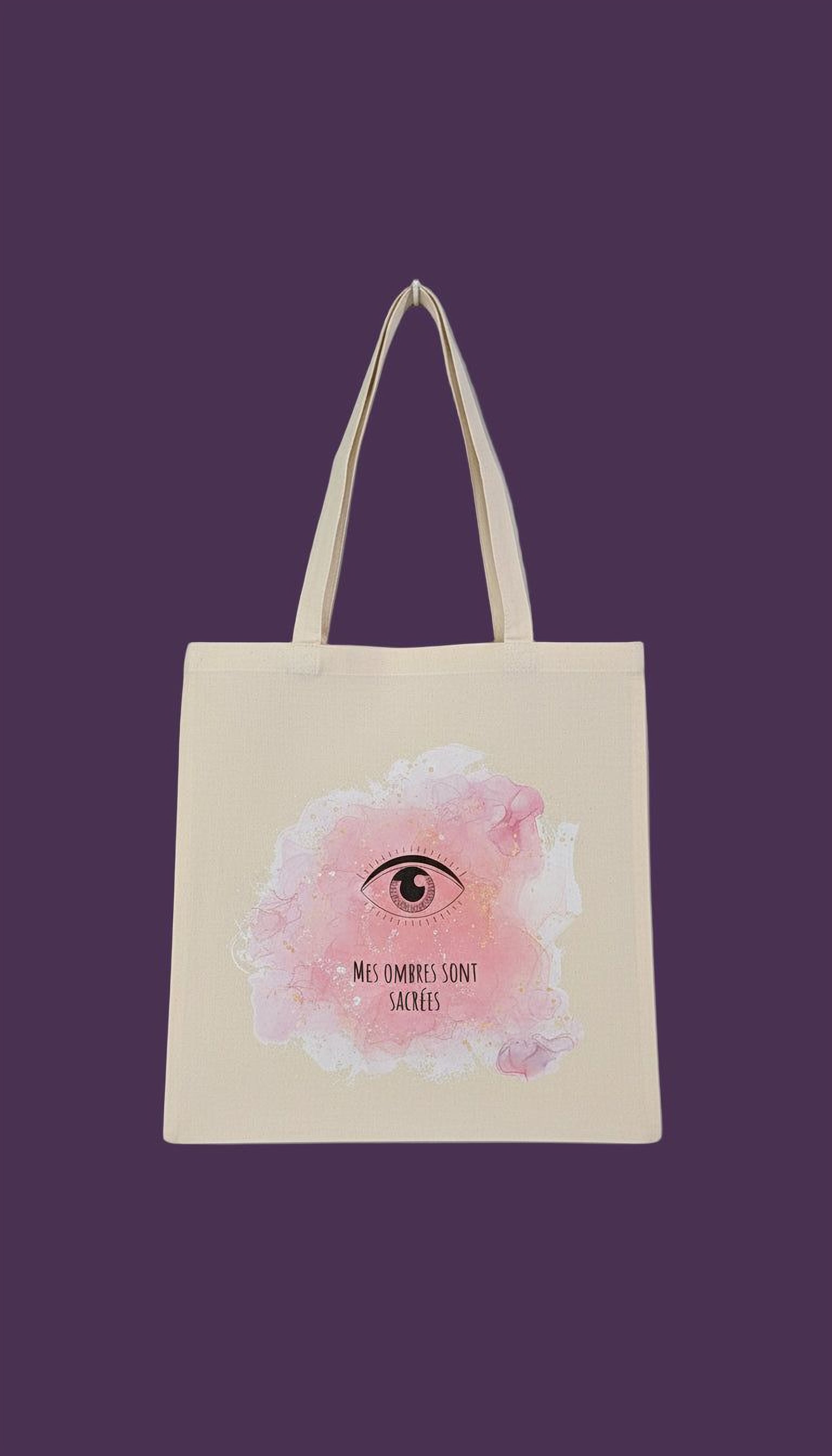 A beige tote bag with a pink eye design and the text "Mes ombre sont sacre" printed on it.
