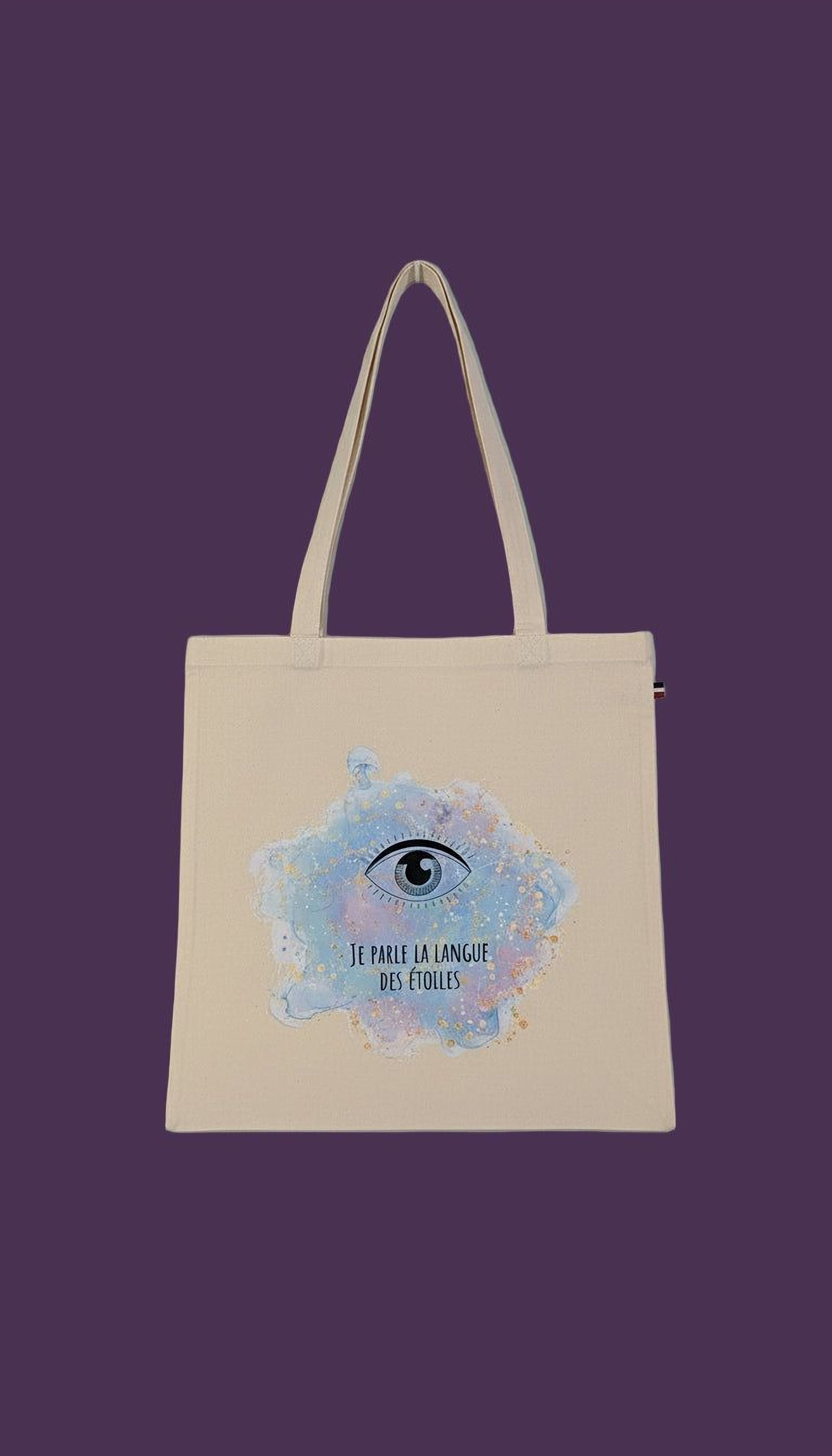 A beige tote bag with a printed design on it, featuring an eye and the text "JE PARLE LA LANGUE DES ETOLLES" (The language of the eye).