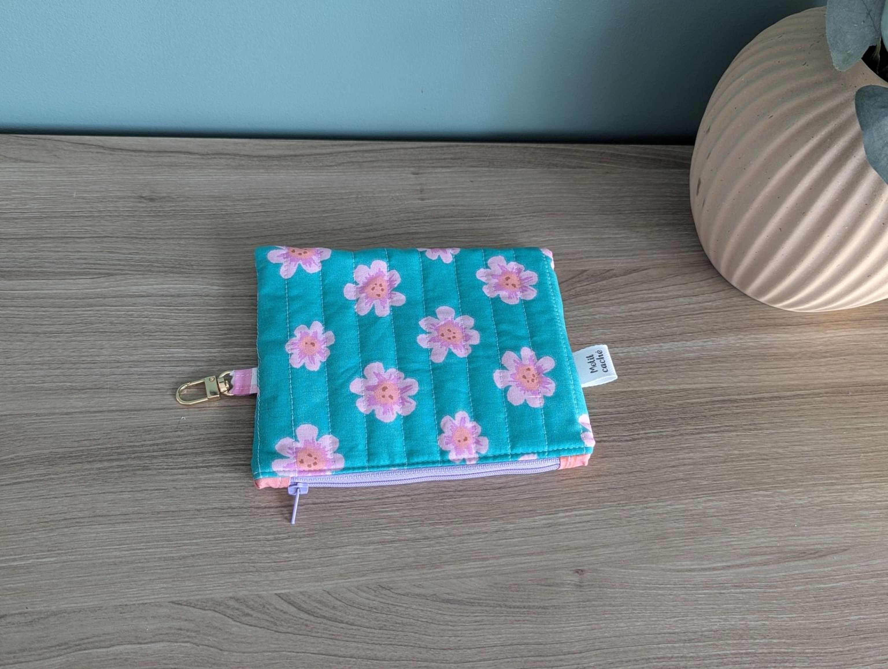 A small, colorful quilted pouch with pink flowers is placed on a wooden surface next to a vase.