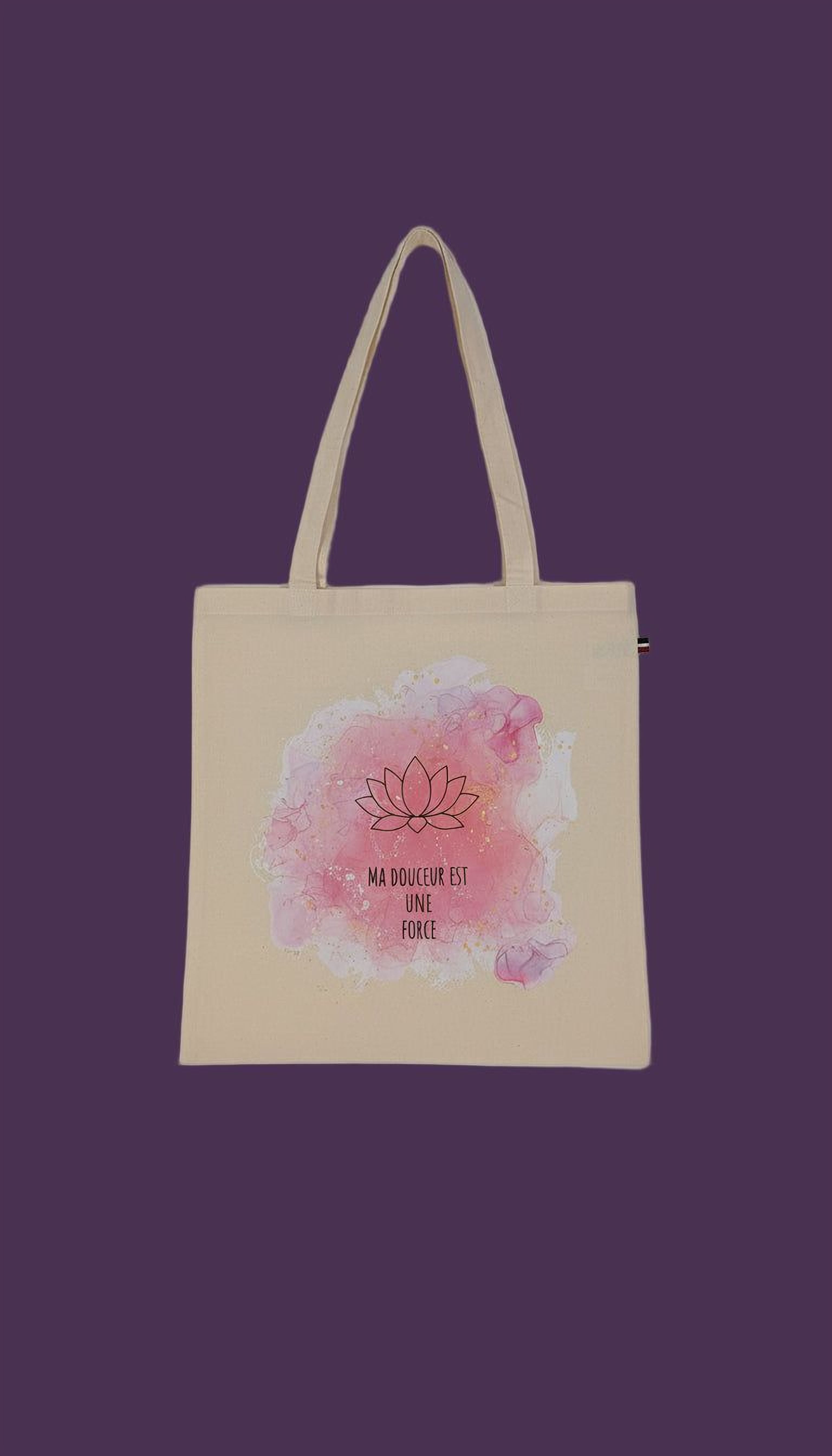 A beige tote bag with a pink and white watercolor design featuring a lotus flower and the text "MA DOUCER EST UNE FORCE" (which means "I am stronger than my circumstances") printed on it.