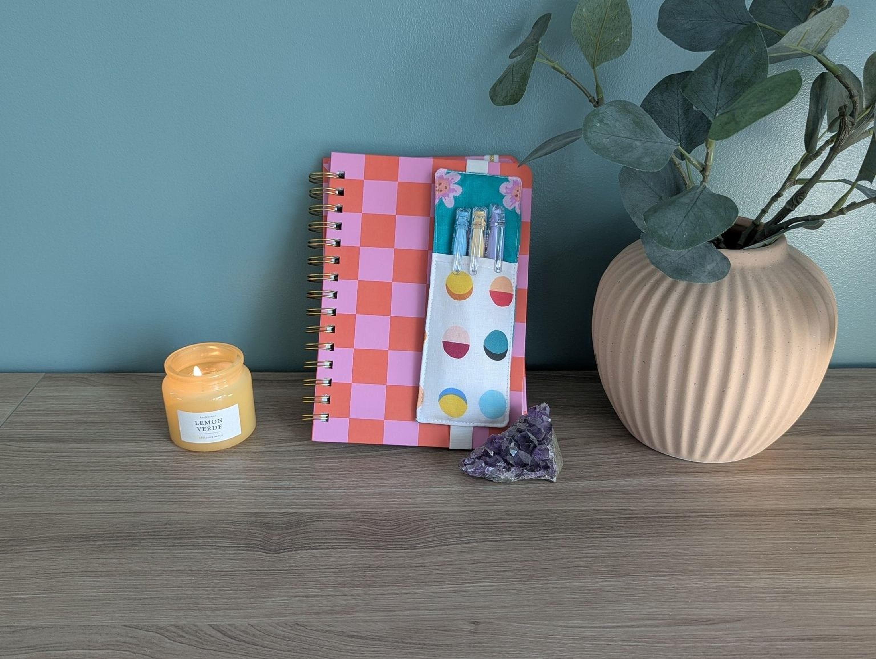 a wooden desk with a pink and orange checkered notebook, a candle, a small crystal, and a plant in a vase.