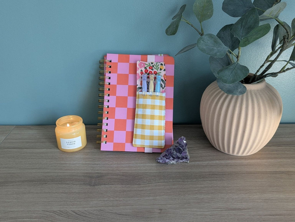 a pink and orange checkered notebook, a candle, a small crystal, and a plant in a vase on a wooden surface.