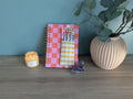 a pink and orange checkered notebook, a candle, a small crystal, and a plant in a vase on a wooden surface.