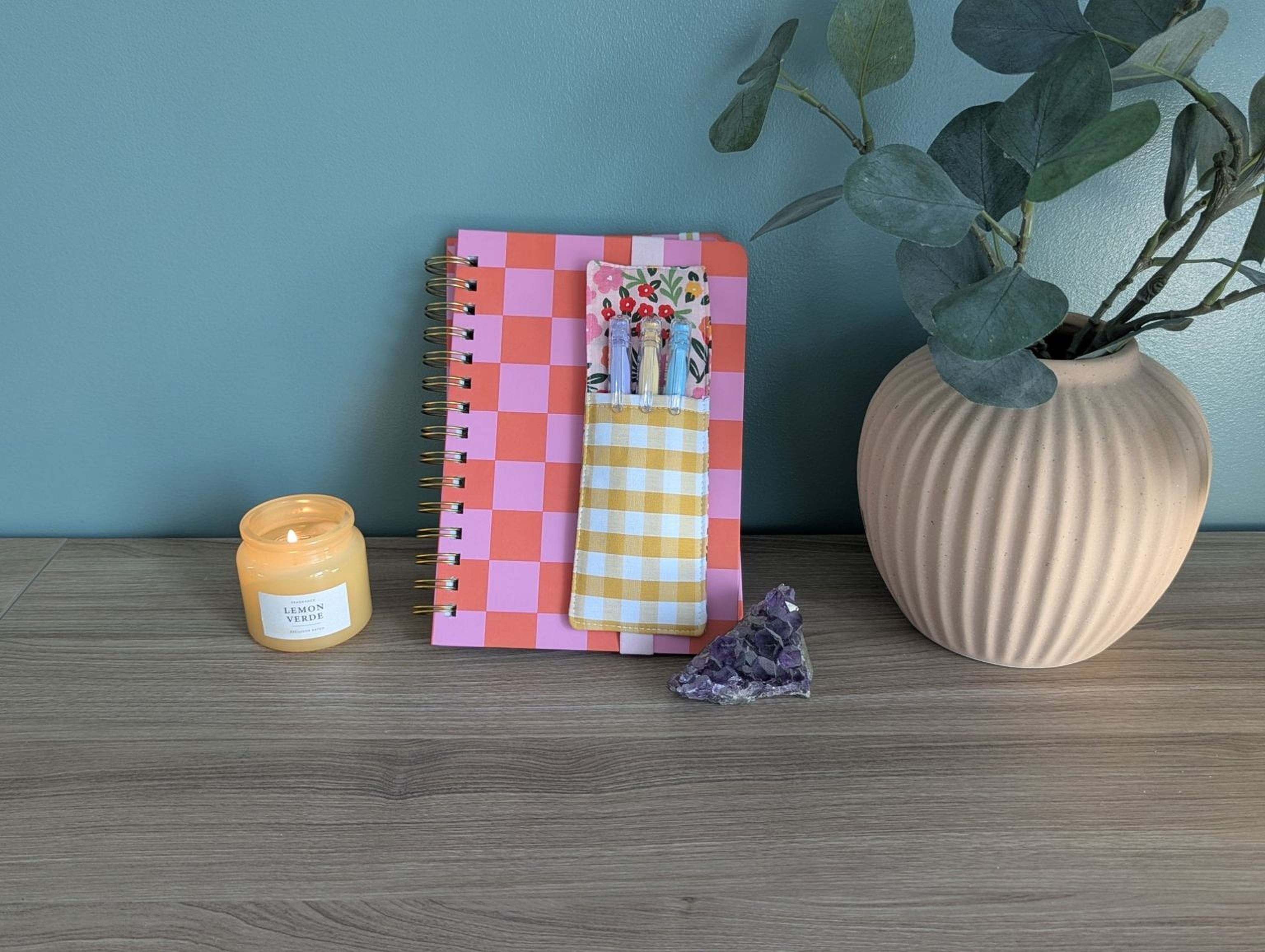 a pink and orange checkered notebook, a candle, a small crystal, and a plant in a vase on a wooden surface.