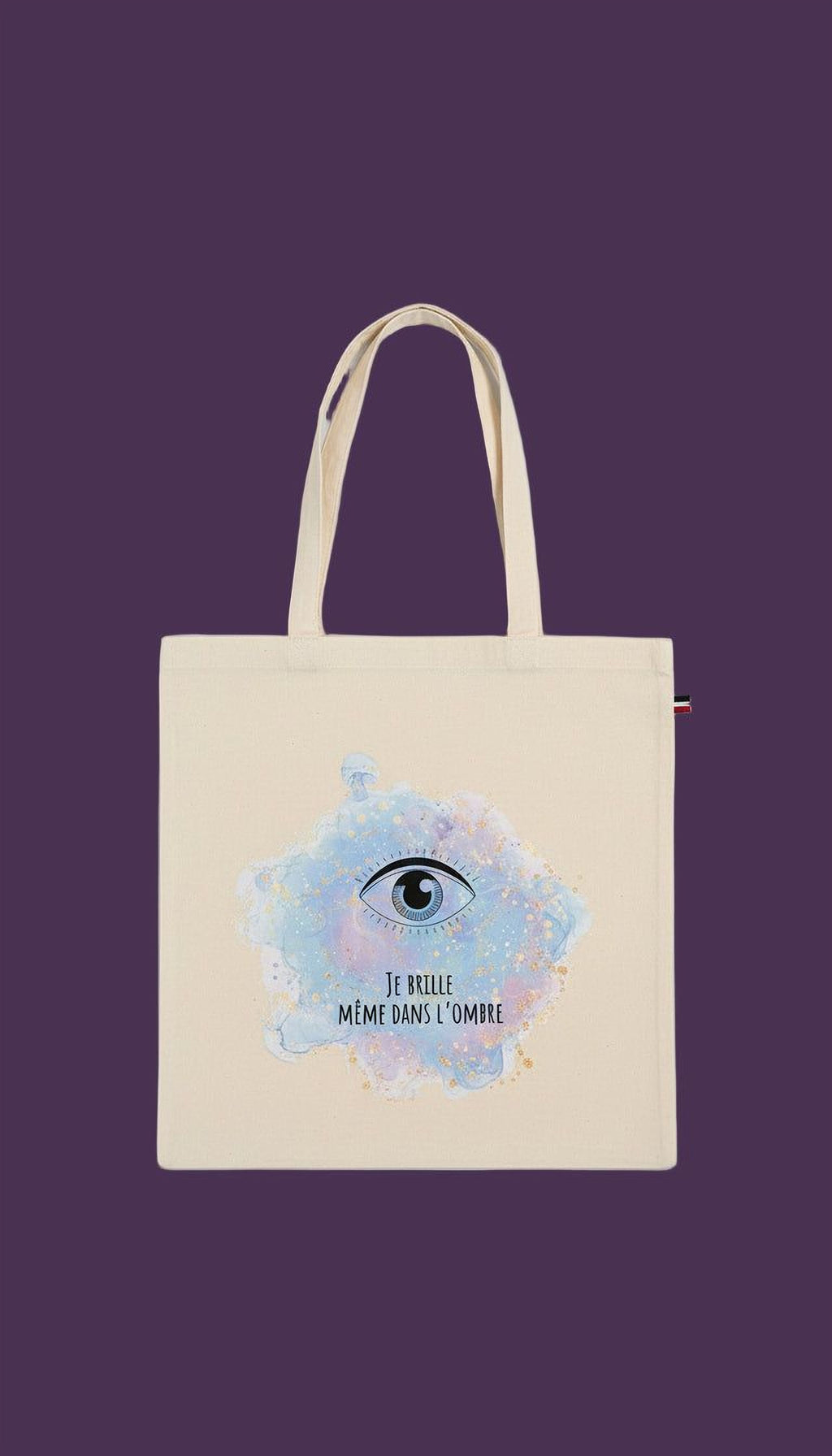 A beige tote bag with a printed design of an eye and the text "J'BLE ME MÉ DANS L'OMBIRE" (The eye is blue) printed on it.
