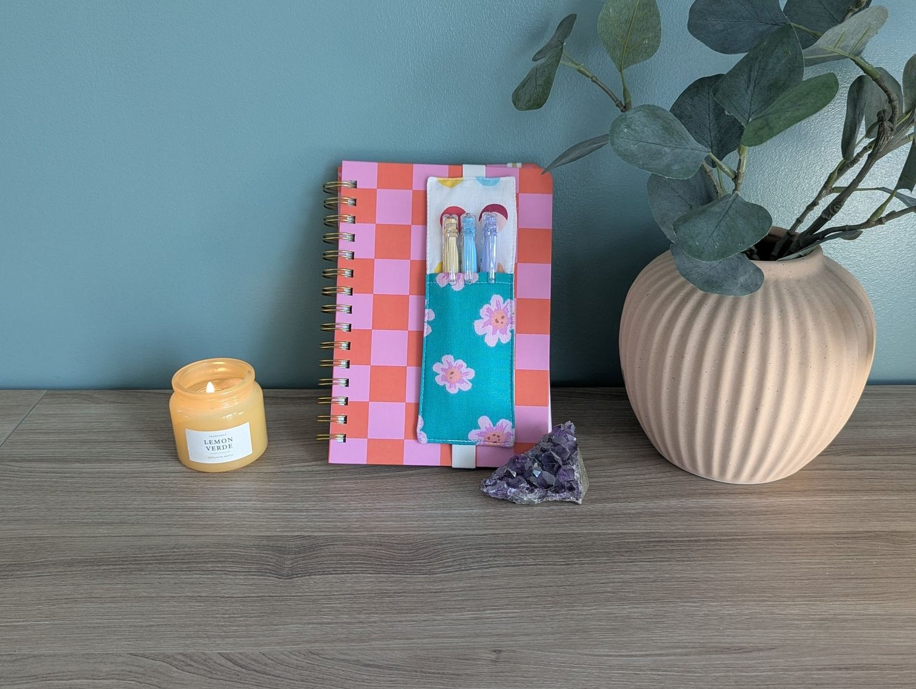 a wooden desk with a pink and orange checkered notebook, a candle, a small crystal, and a plant in a vase.