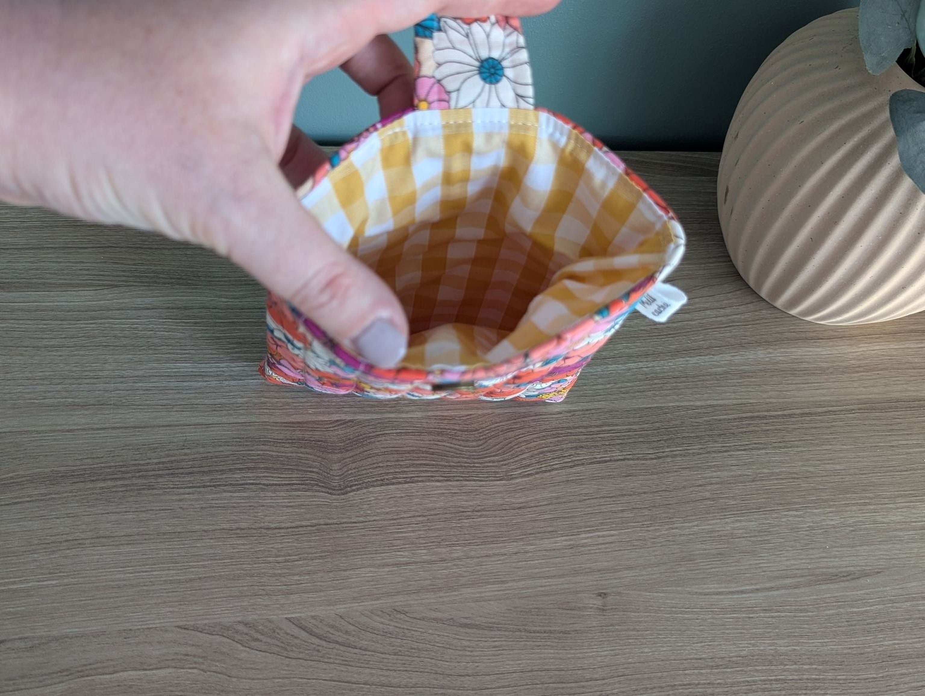 A hand is holding a colorful pouch with a floral pattern, placed on a wooden surface next to a vase.