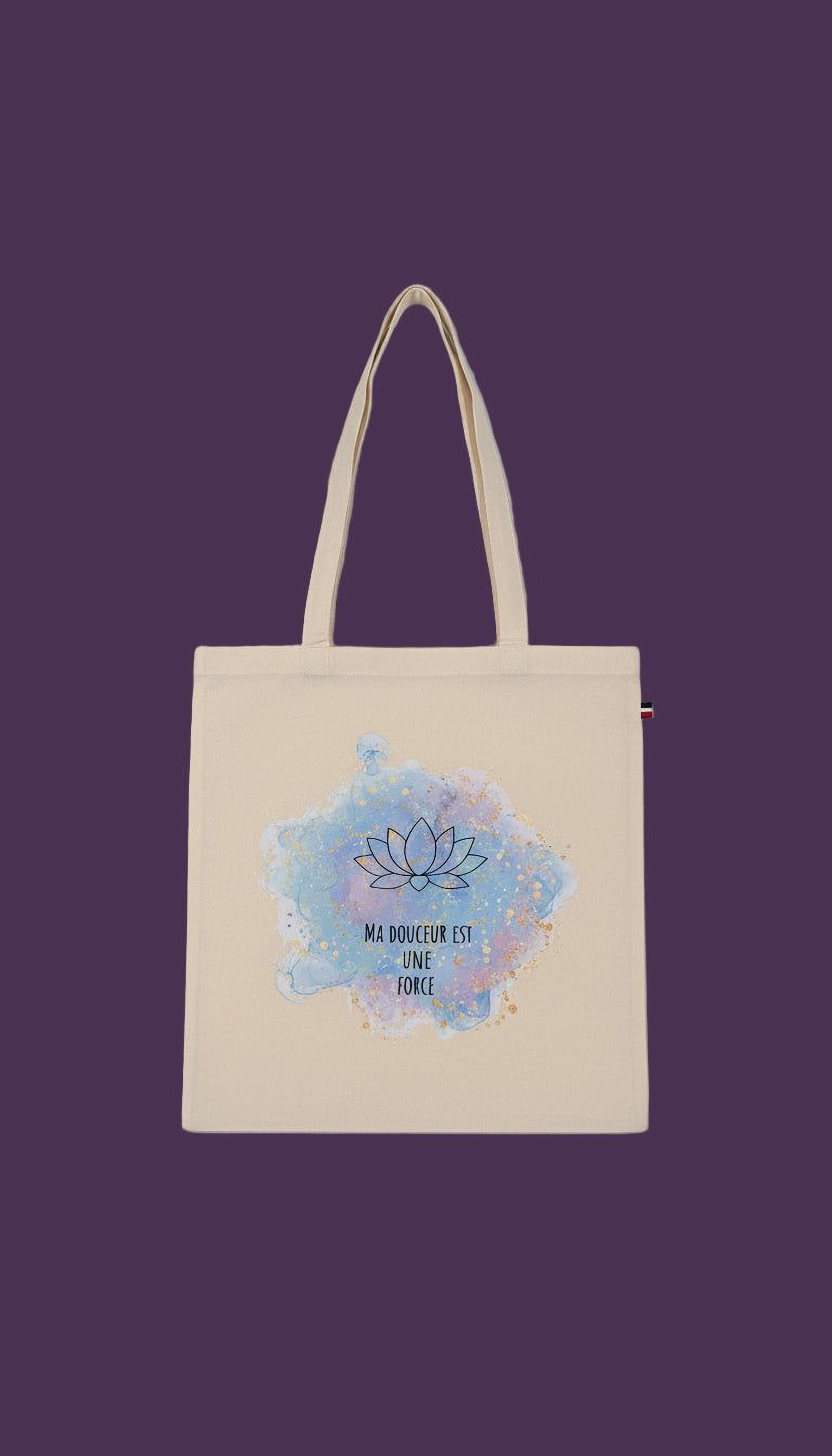 A beige tote bag with a blue and purple watercolor design featuring a lotus flower and the text "MA DOUCEUR EST MÈS FÈRCE" (which means "The flower is the force" in French).