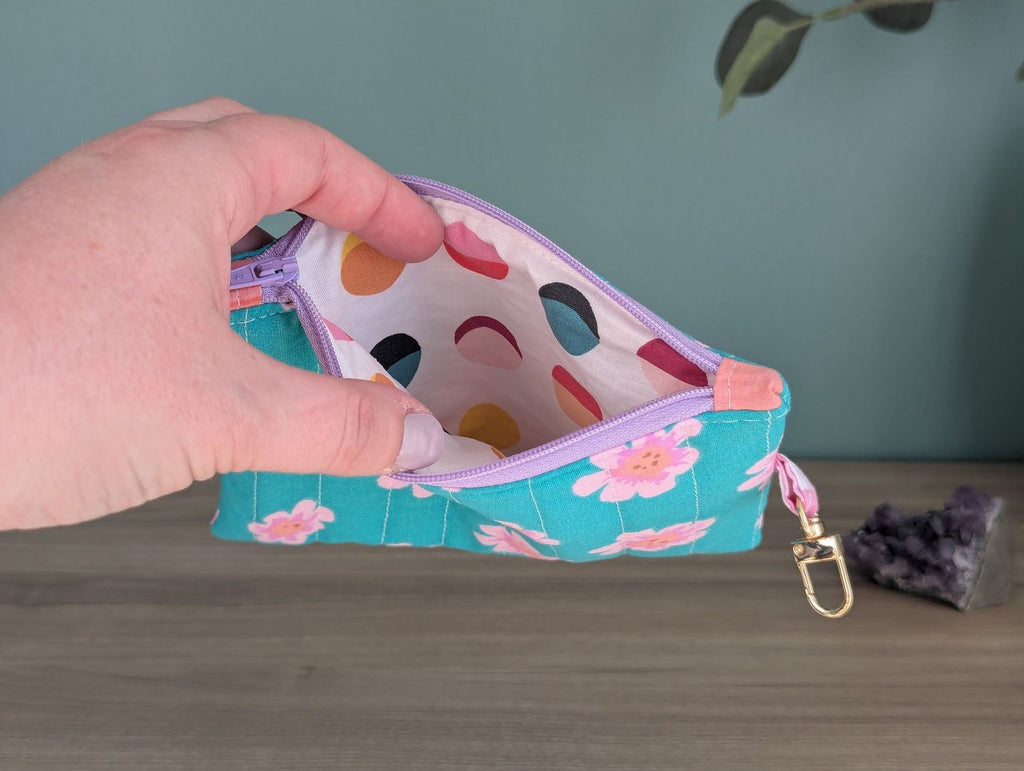 A hand is holding a colorful polka dot fabric pouch with a floral pattern, placed on a wooden surface next to a small crystal or gemstone.