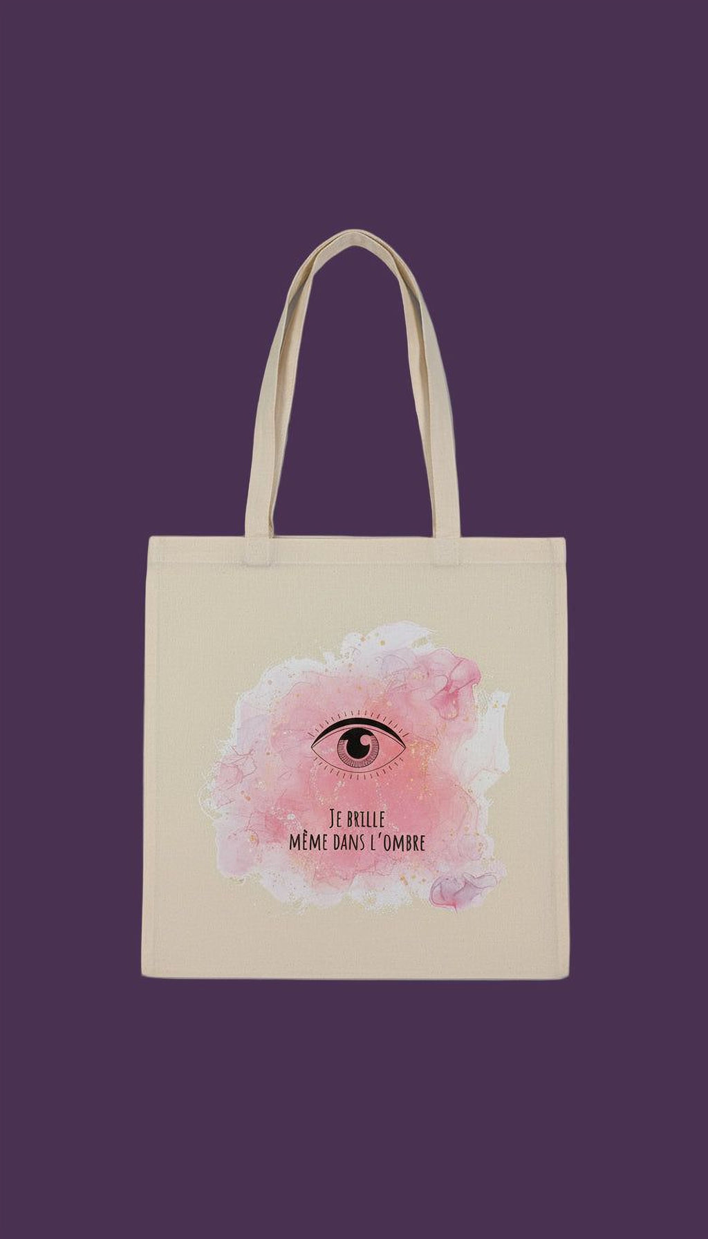 A beige tote bag with a pink eye design and the text "Te brille, me meme dans l'ombre" printed on it.