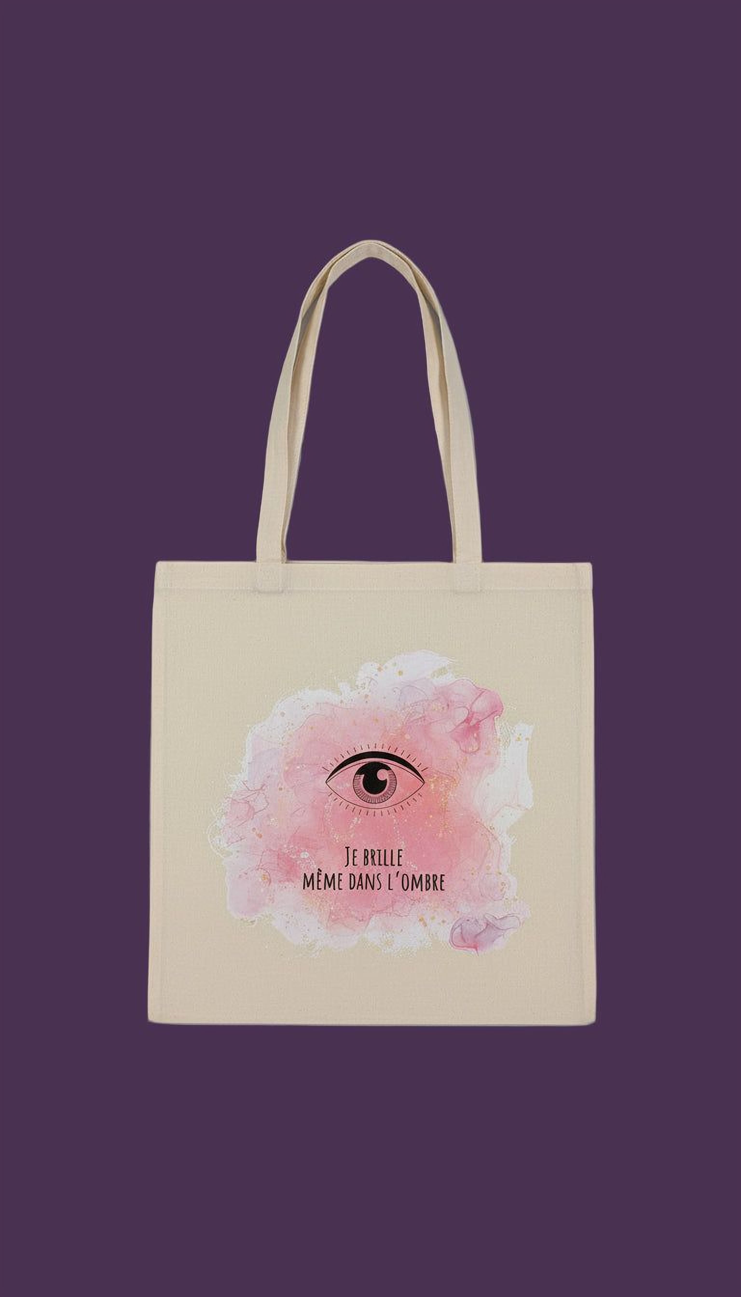 A beige tote bag with a pink eye design and the text "Te brille, me meme dans l'ombre" printed on it.