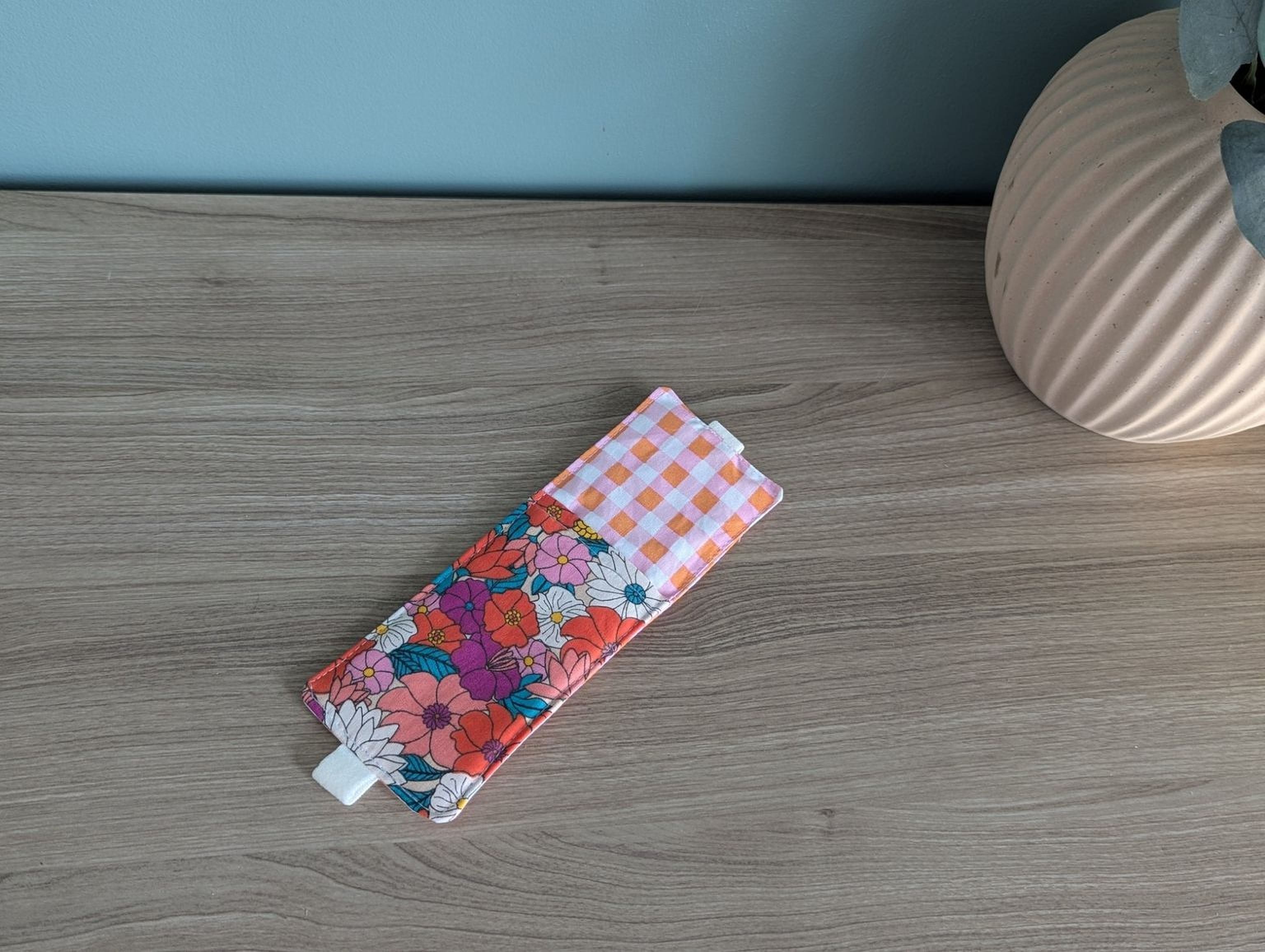 A colorful floral patterned case or wallet is placed on a wooden surface next to a vase.