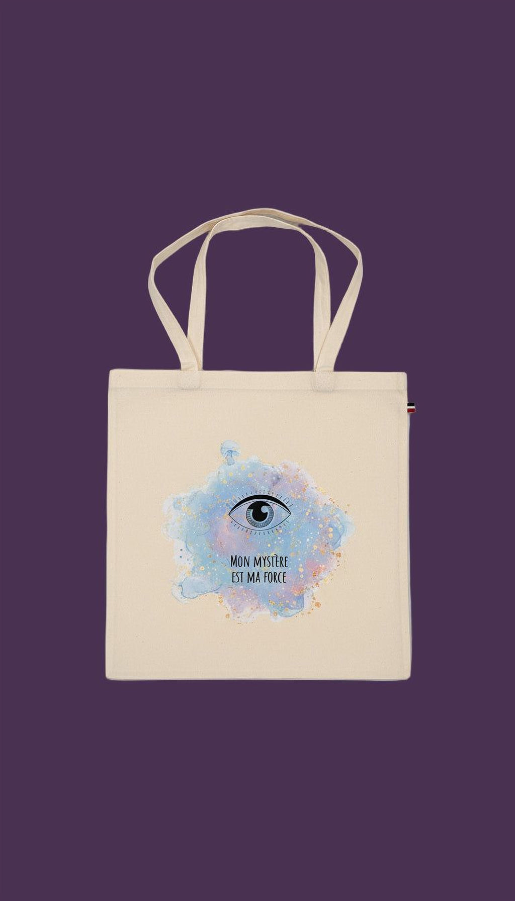 A beige tote bag with a printed design on it, featuring an eye and the text "MON MYSTÉRE EST MA FORCE" (My mystery is greater than my force).