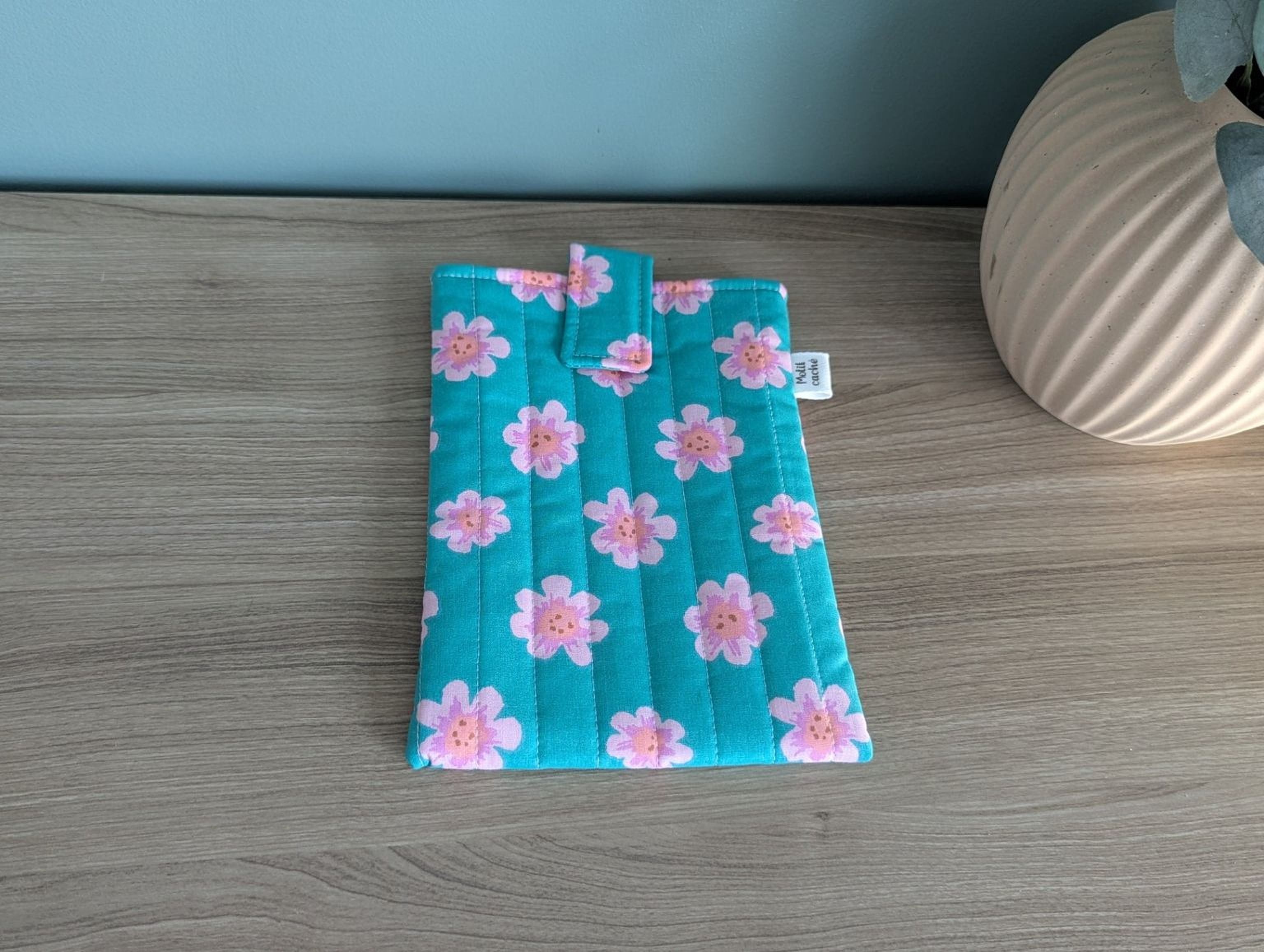A teal-colored fabric pouch with a floral pattern is placed on a wooden surface next to a white vase.