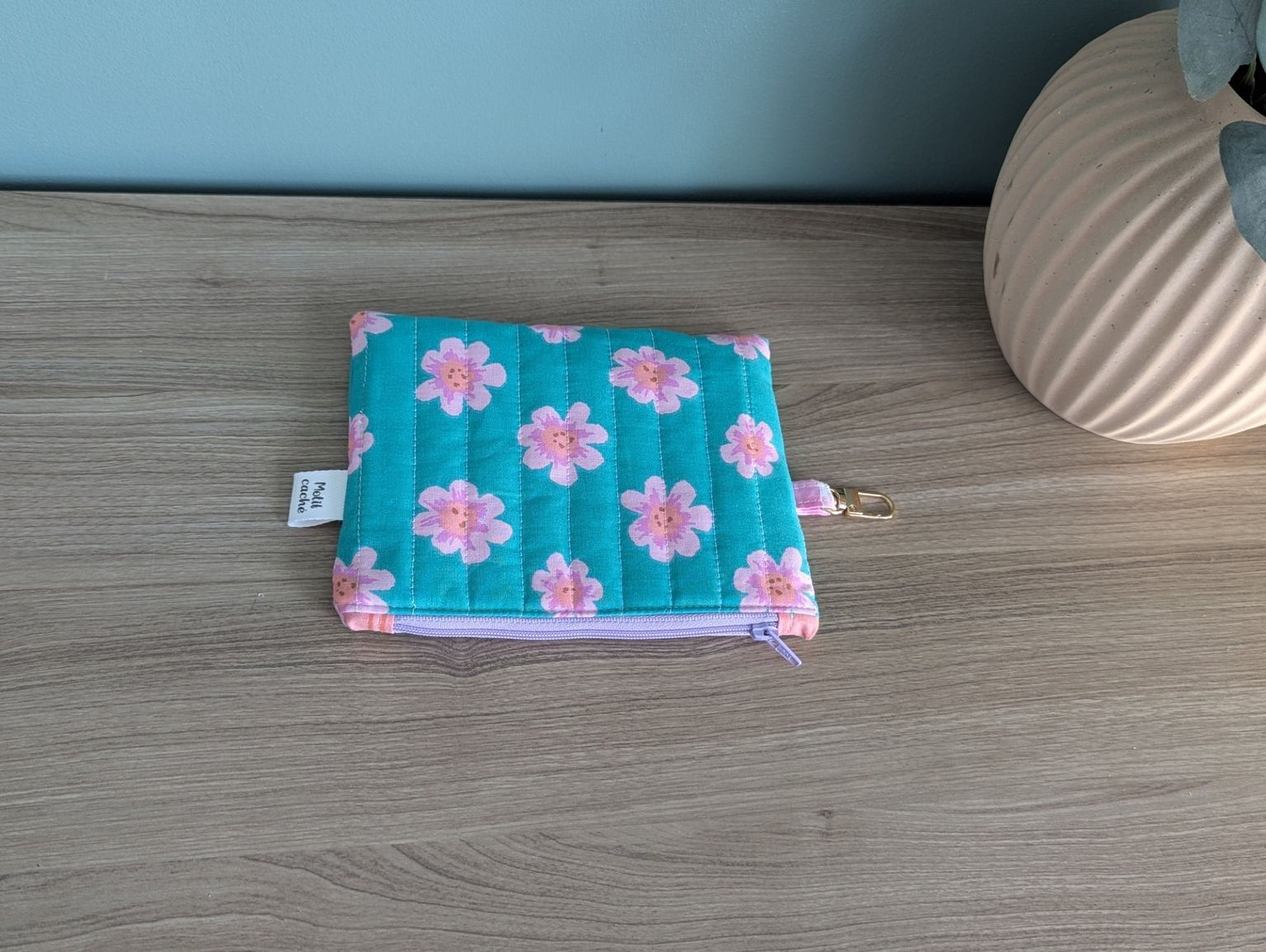 A small, square-shaped, teal-colored pouch with pink flowers printed on it, placed on a wooden surface next to a white vase.