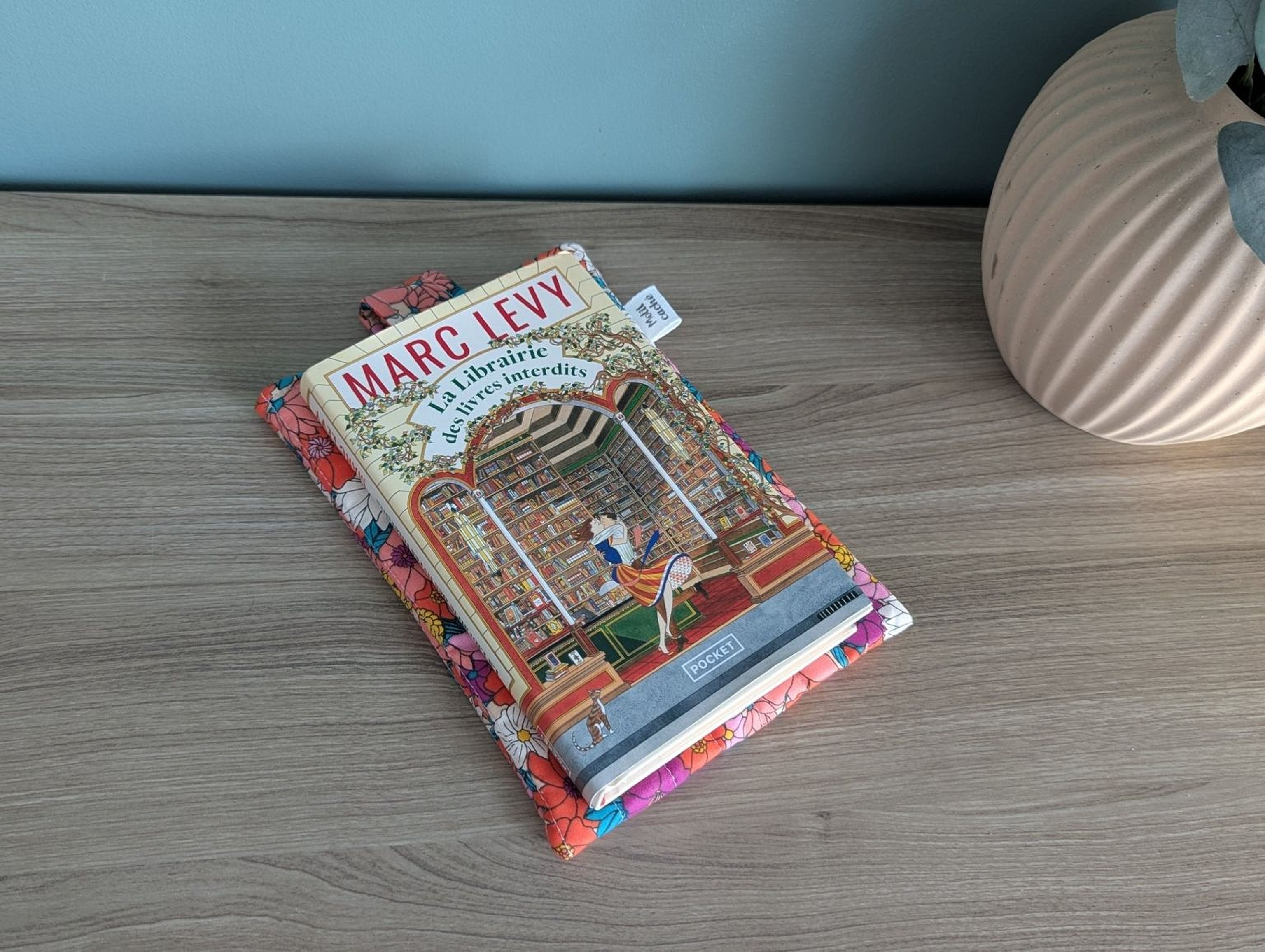 A book titled "Marc Levy" is placed on a wooden surface, with a colorful book cover featuring an illustration of a library scene.