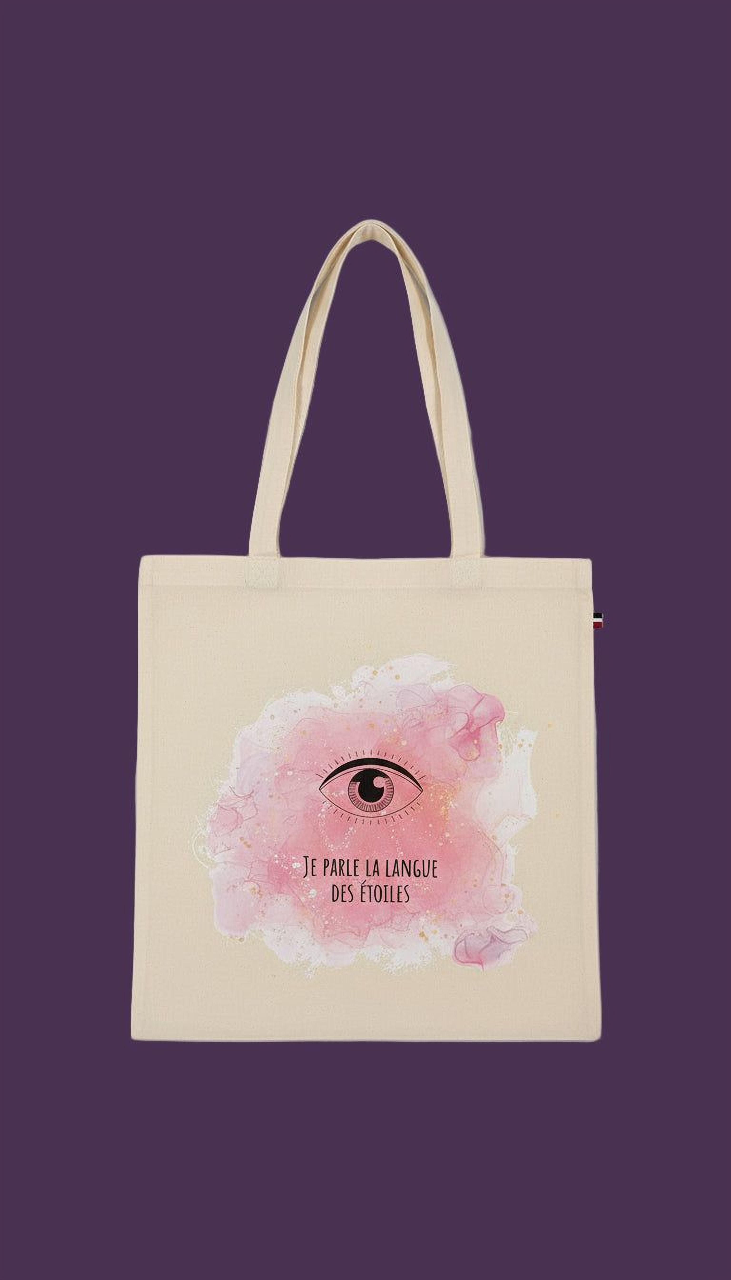 A beige tote bag with a pink and white watercolor design featuring an eye and the text "JE PARLE LA LANGUE DES FOULS" (The language is French).