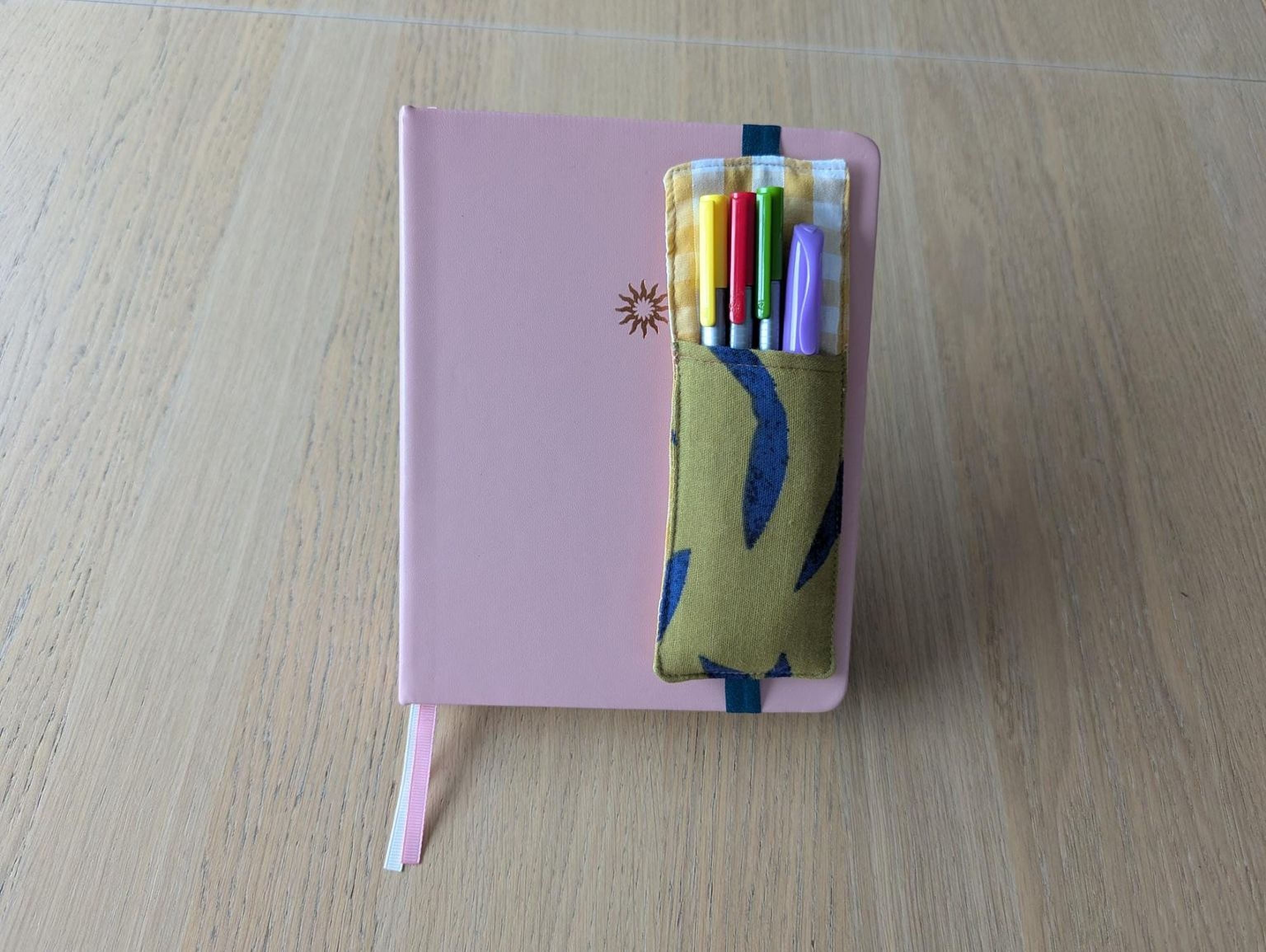 A pink notebook with a pen holder attached to it, containing a variety of colorful pens.