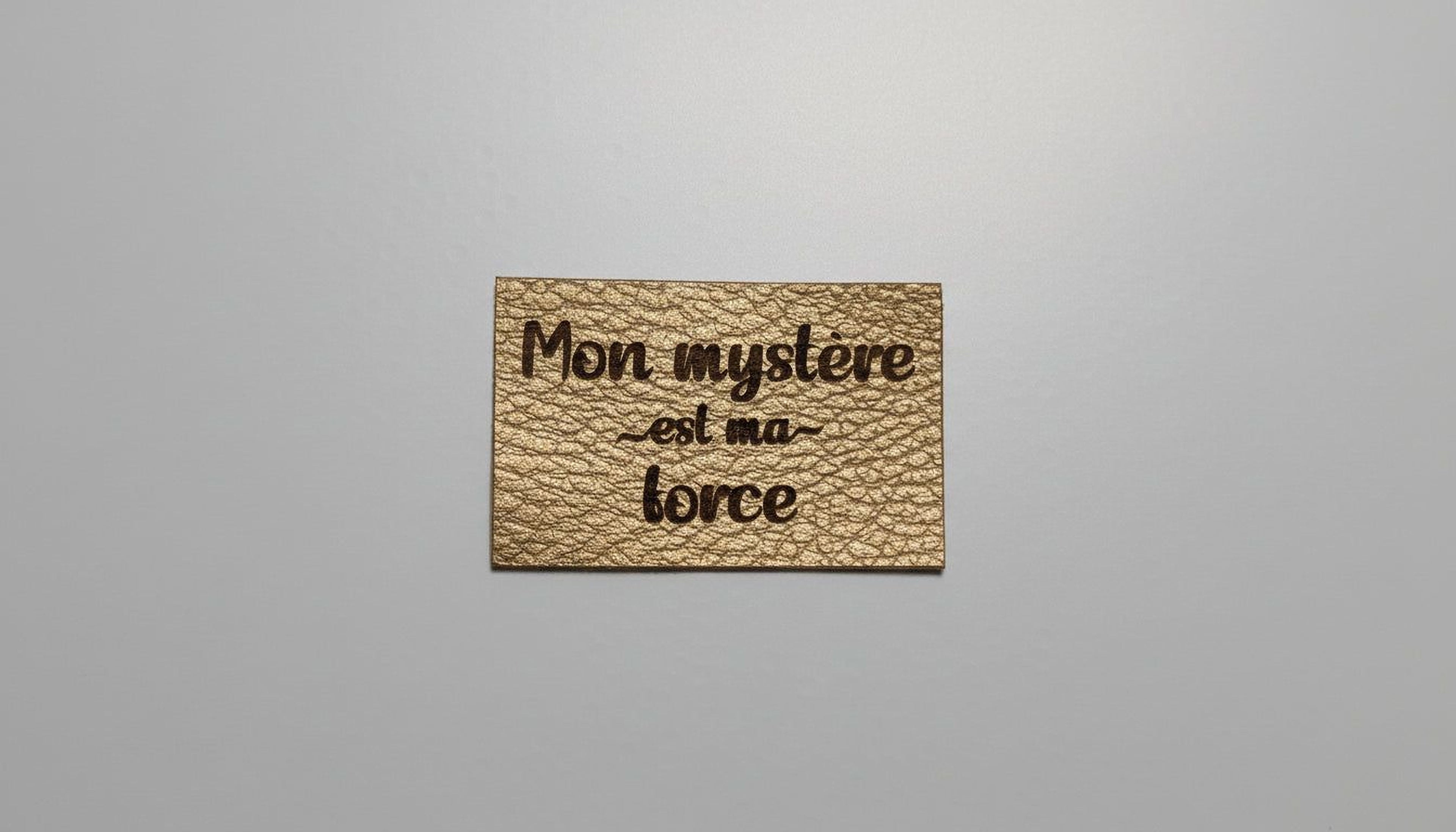 a rectangular piece of leather with the text "Mon mystere est ma force" printed on it.