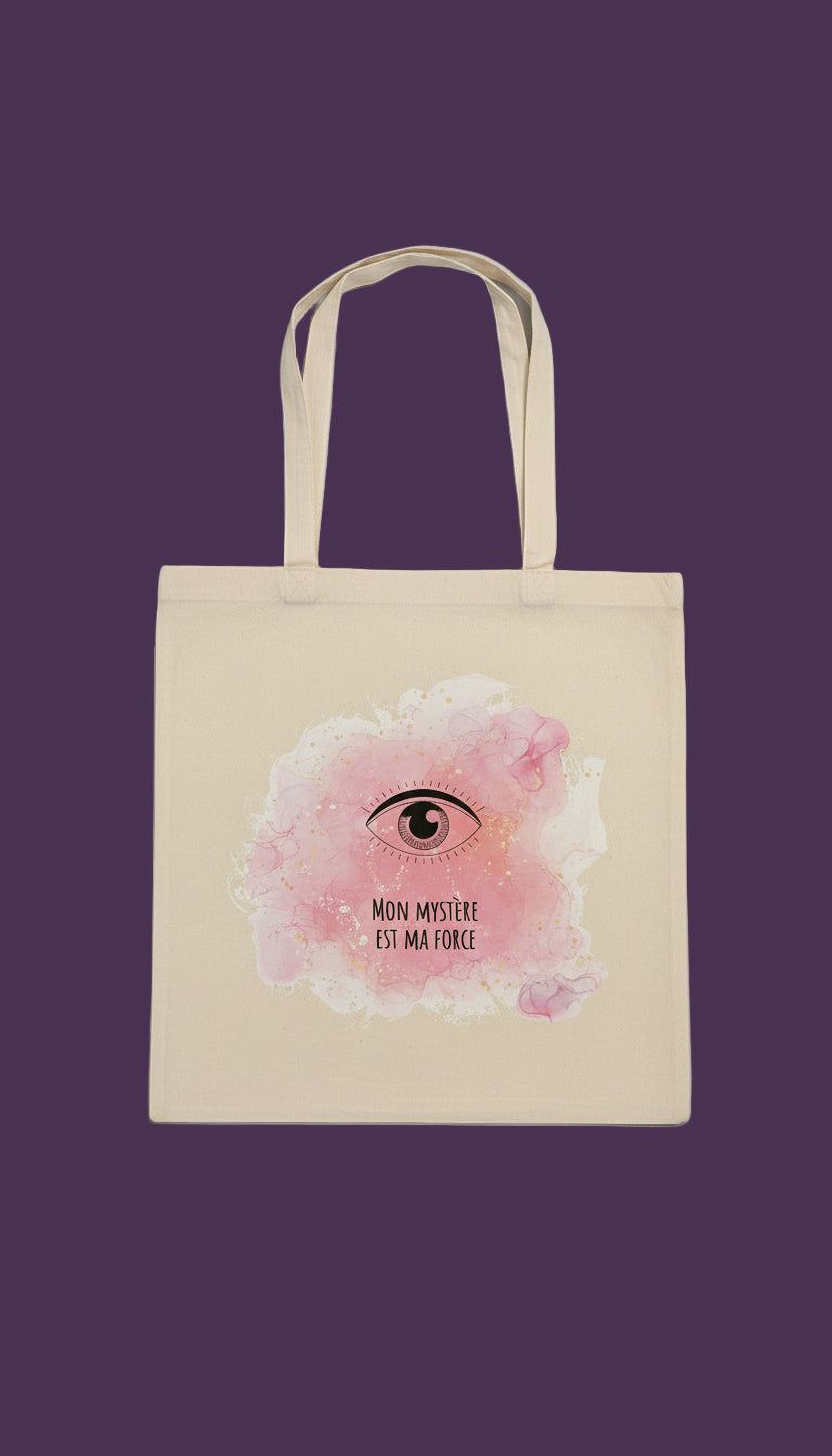 A beige tote bag with a pink eye design and the text "Mon mystere est ma force" printed on it.