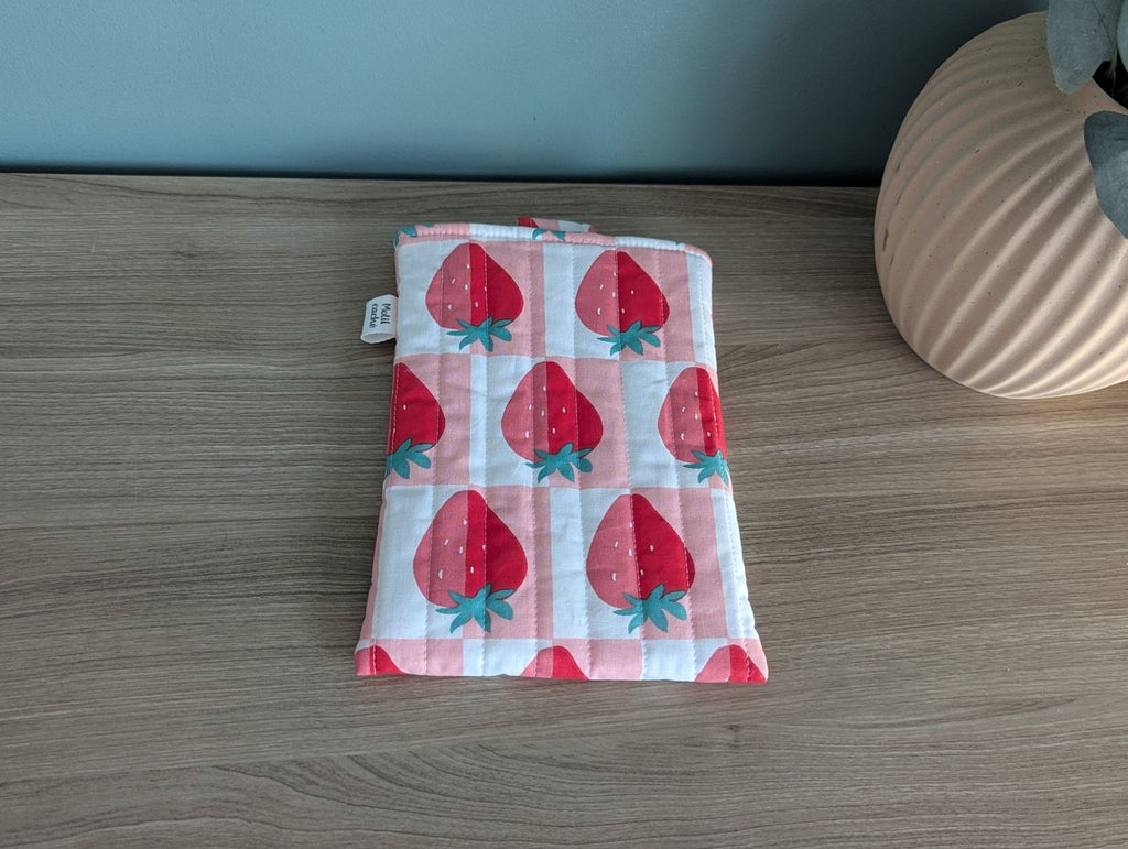A quilted pouch with a strawberry pattern is placed on a wooden surface next to a vase.