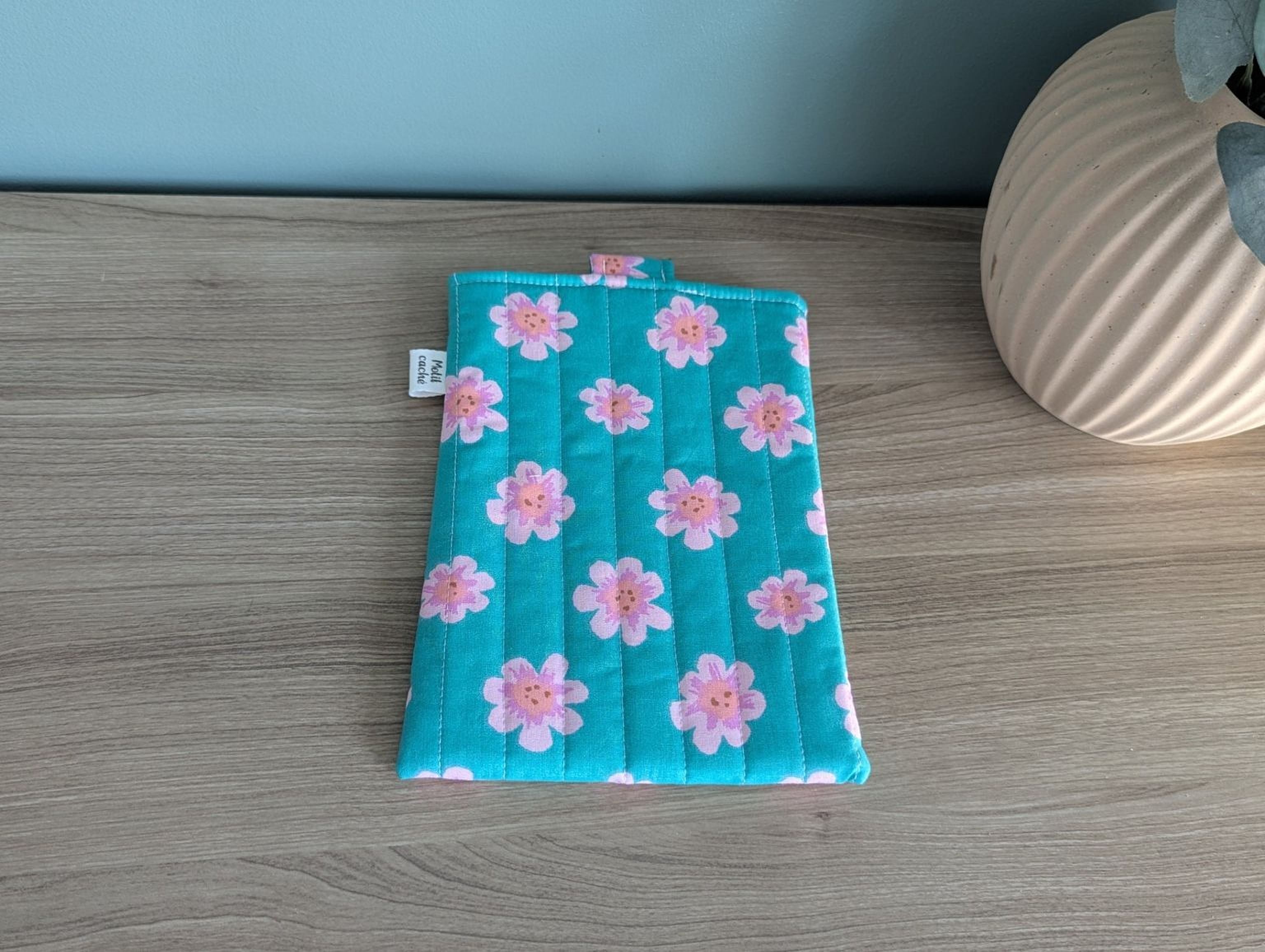A teal-colored quilted pouch with a pattern of pink flowers is placed on a wooden surface next to a white vase.