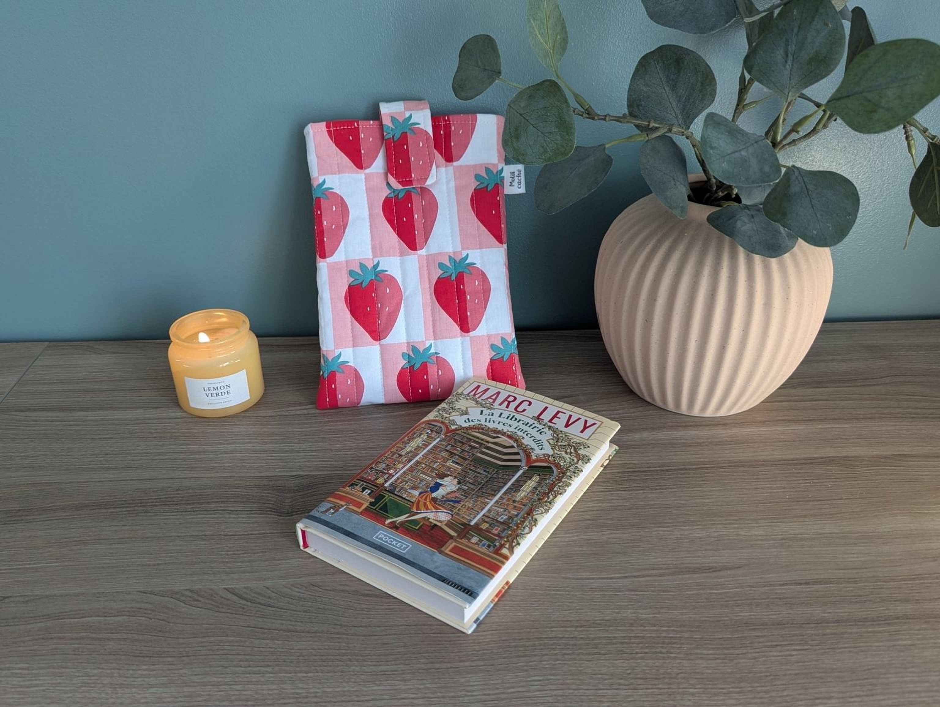 a wooden table with a book, a candle, and a plant. The book is titled "MARGOLLE VVERD" and is placed on the table. The candle is positioned to the left of the book, and the plant is located to the right of the candle.