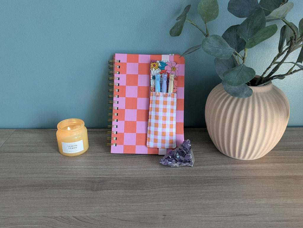 a pink and orange checkered notebook, a candle, a small crystal, and a beige vase with a plant on a wooden surface.