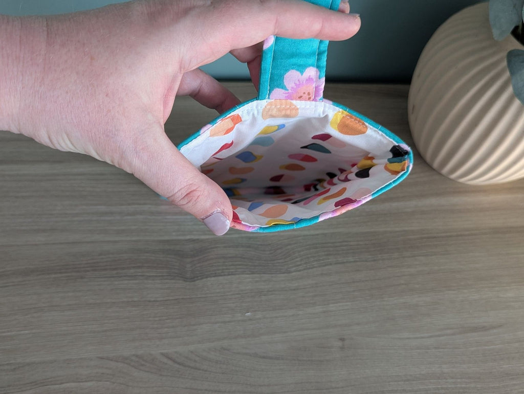 A hand holding a colorful, patterned purse with a teal handle, placed on a wooden surface.