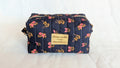 A black and pink floral patterned cosmetic bag with a gold-colored label that says "Chaque souille est un renaise"
