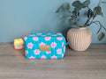 A blue floral makeup bag with a gold zipper detail sits on a wooden surface, accompanied by a lit candle and a vase with green leaves.
