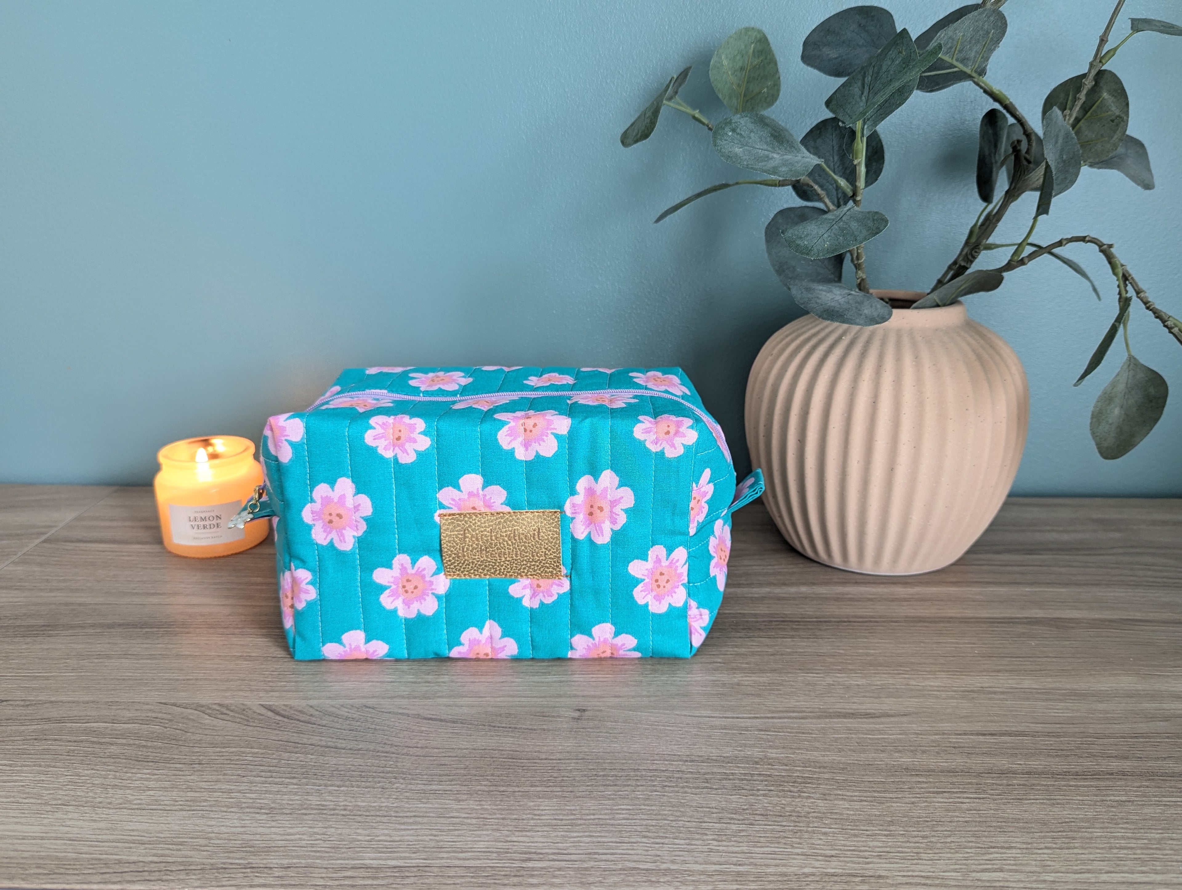 A blue floral makeup bag with a gold zipper detail sits on a wooden surface, accompanied by a lit candle and a vase with green leaves.