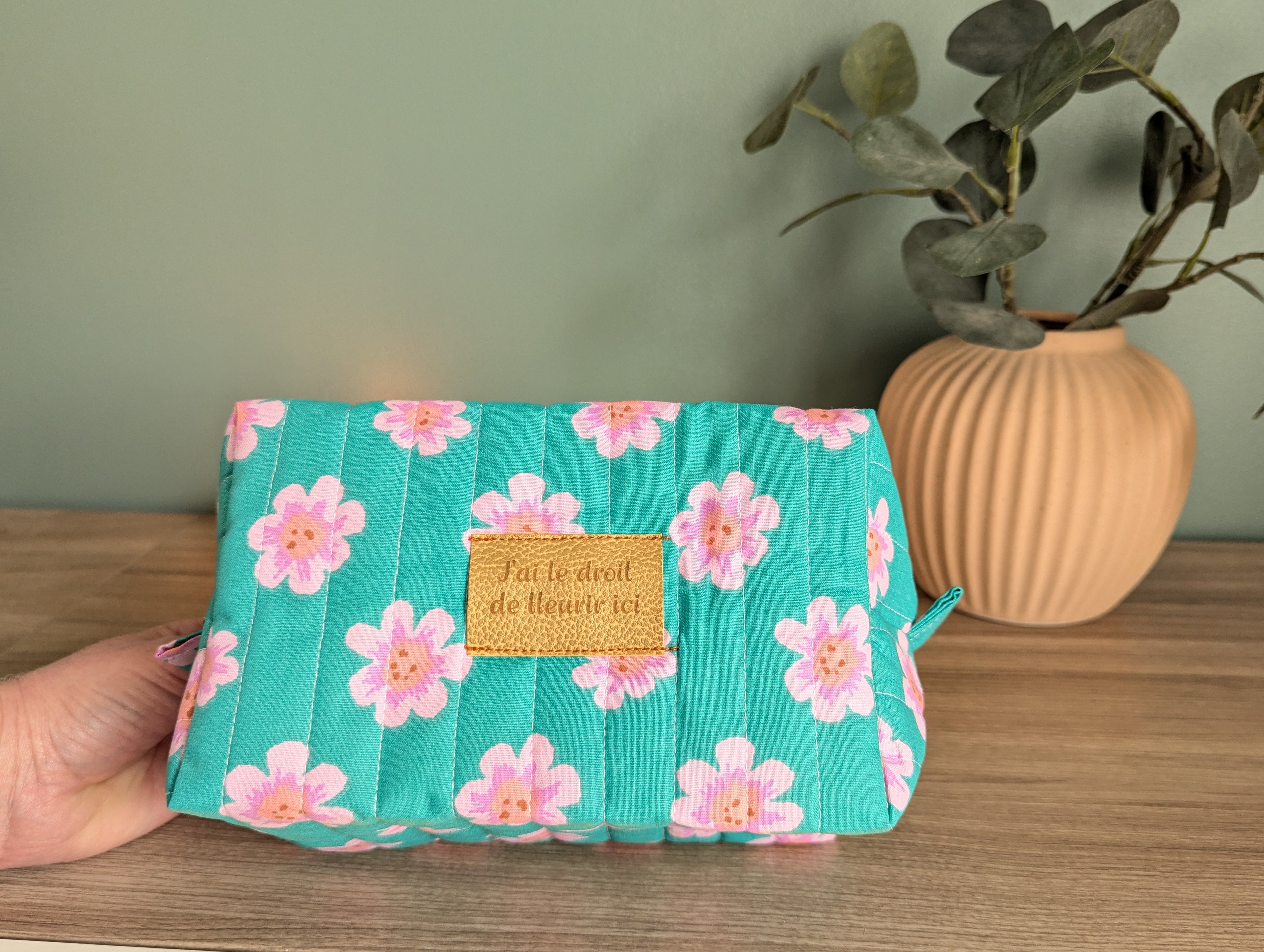 A hand holding a teal-colored makeup bag with a floral pattern, placed on a wooden surface next to a potted plant.