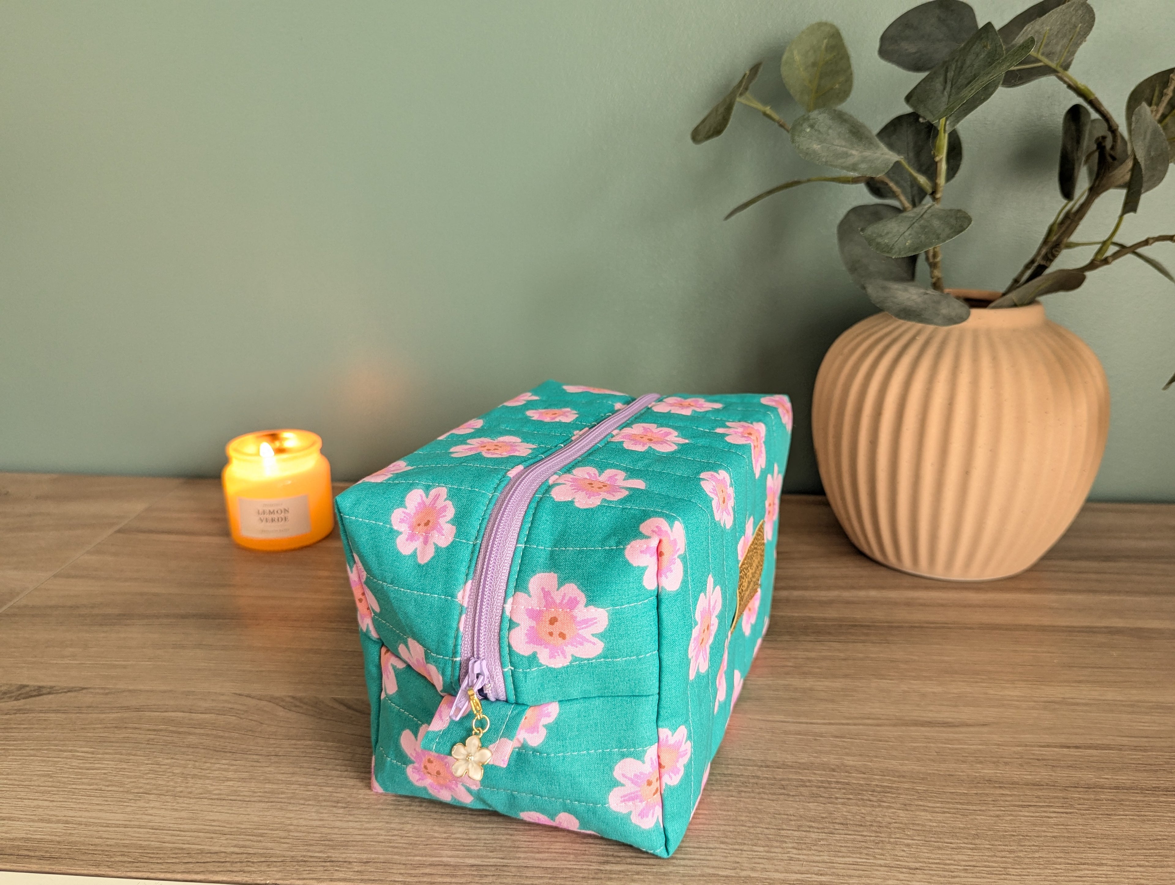 A teal floral makeup bag with a purple zipper sits on a wooden surface, accompanied by a small orange candle and a potted plant.