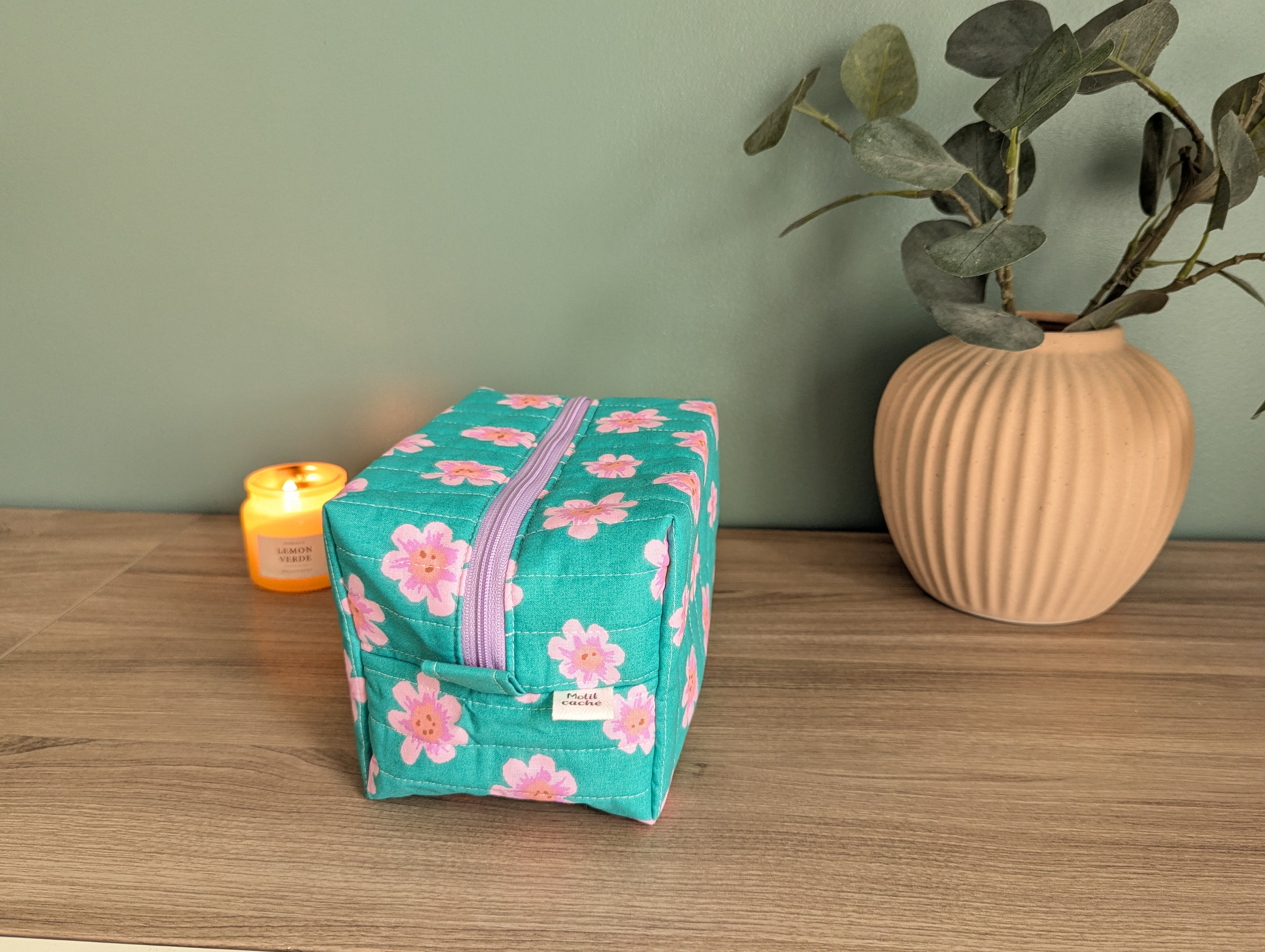 A green floral-patterned bag sits on a wooden surface next to a beige vase with greenery, with a lit candle in the background.