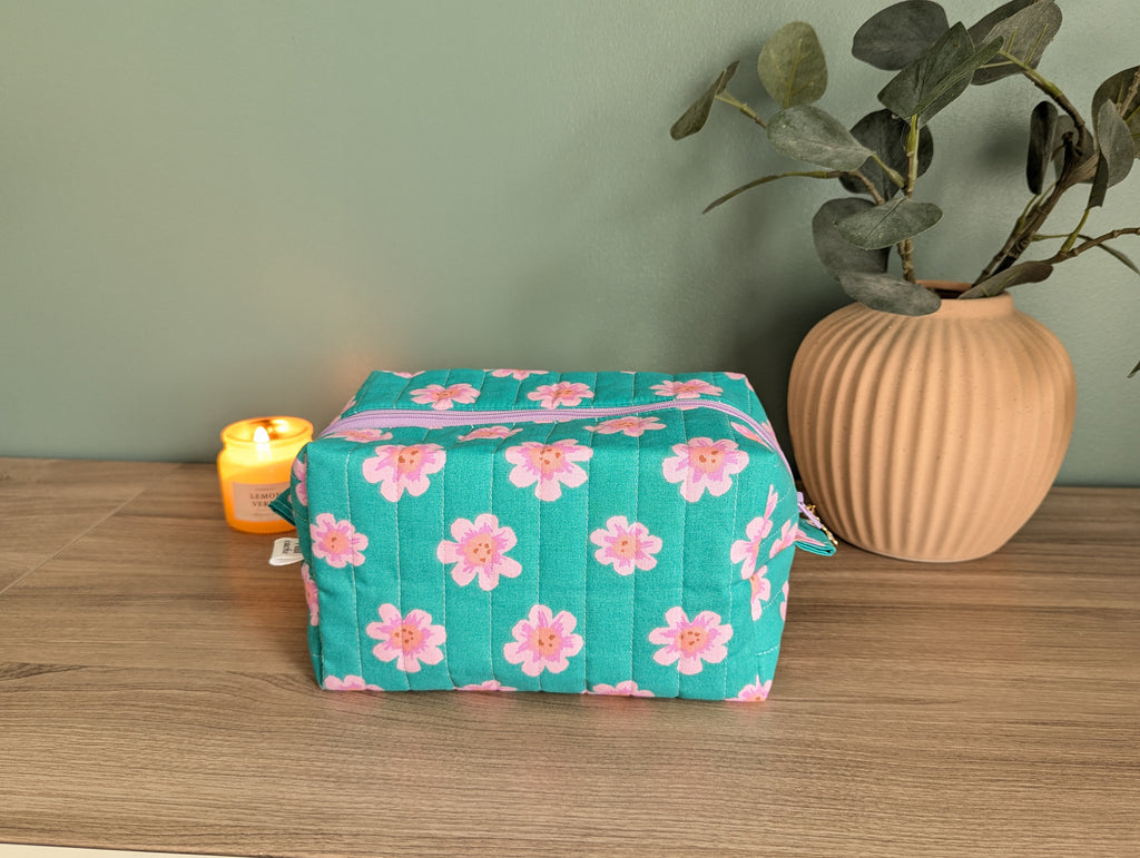 A green and pink floral makeup bag sits on a wooden surface, accompanied by a small yellow candle and a potted plant.