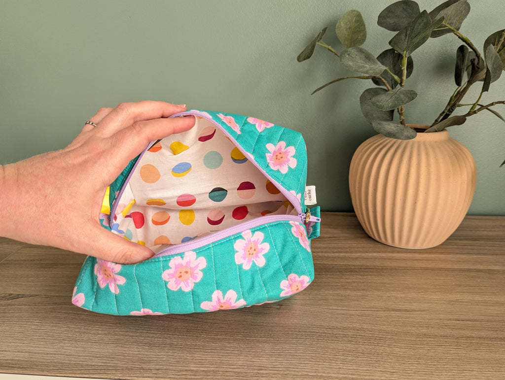 A hand is holding open a colorful polka dot makeup bag on a wooden surface, with a potted plant in the background.