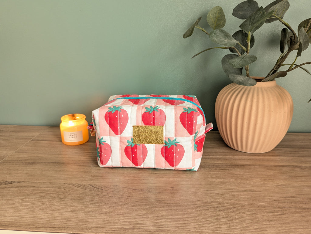 A pink and white cosmetic bag with a strawberry pattern sits on a wooden surface, accompanied by a small yellow candle and a potted plant.