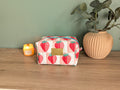 A pink and white cosmetic bag with a strawberry pattern sits on a wooden surface, accompanied by a small yellow candle and a potted plant.