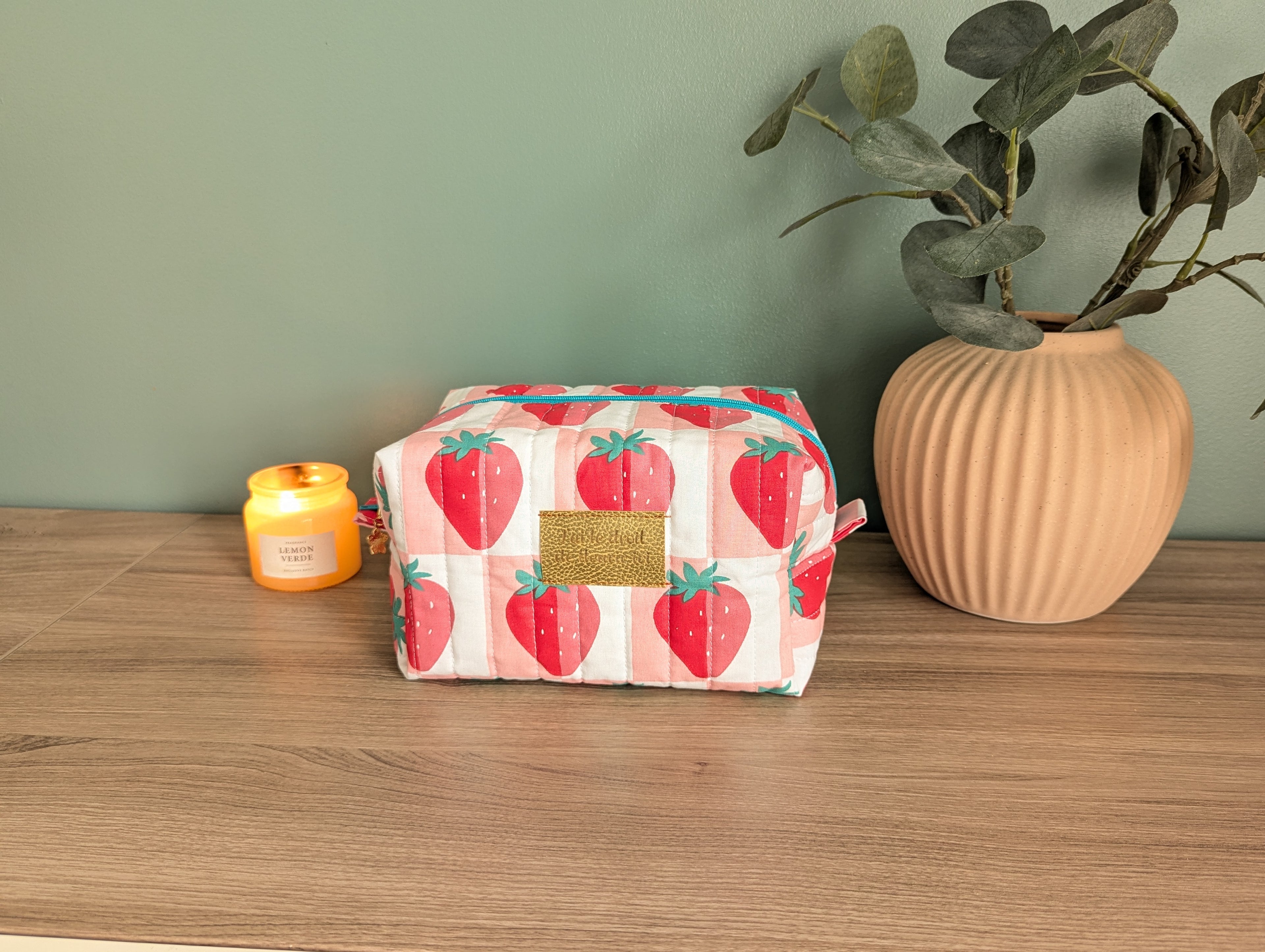 A pink and white cosmetic bag with a strawberry pattern sits on a wooden surface, accompanied by a small yellow candle and a potted plant.