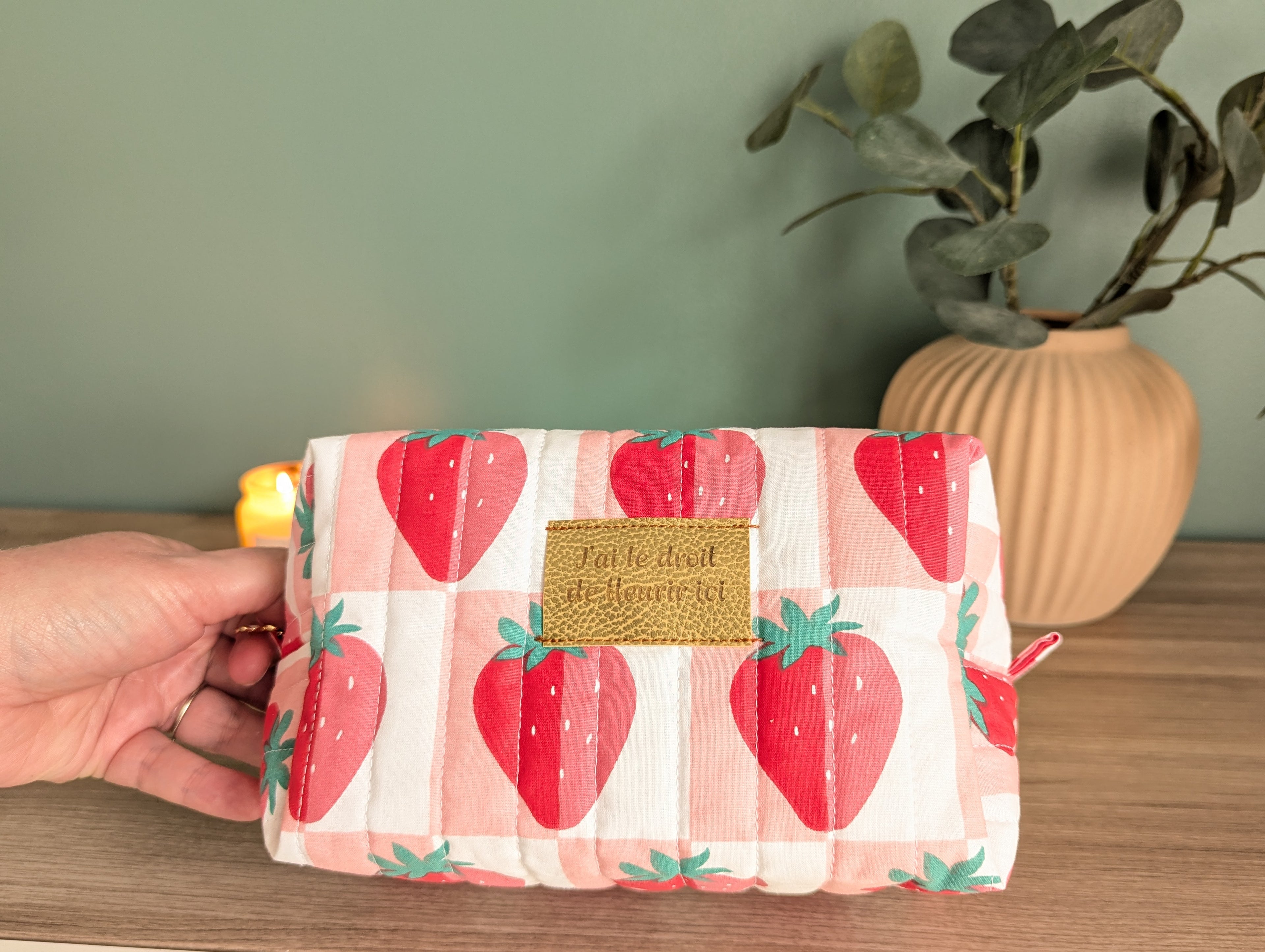 A hand holding a makeup bag with a strawberry pattern, placed on a wooden surface next to a potted plant.