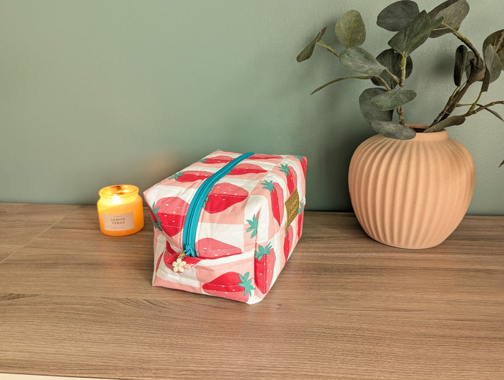 A pink and white striped cosmetic bag with a teal zipper sits on a wooden surface next to a small yellow candle and a beige vase with green leaves.