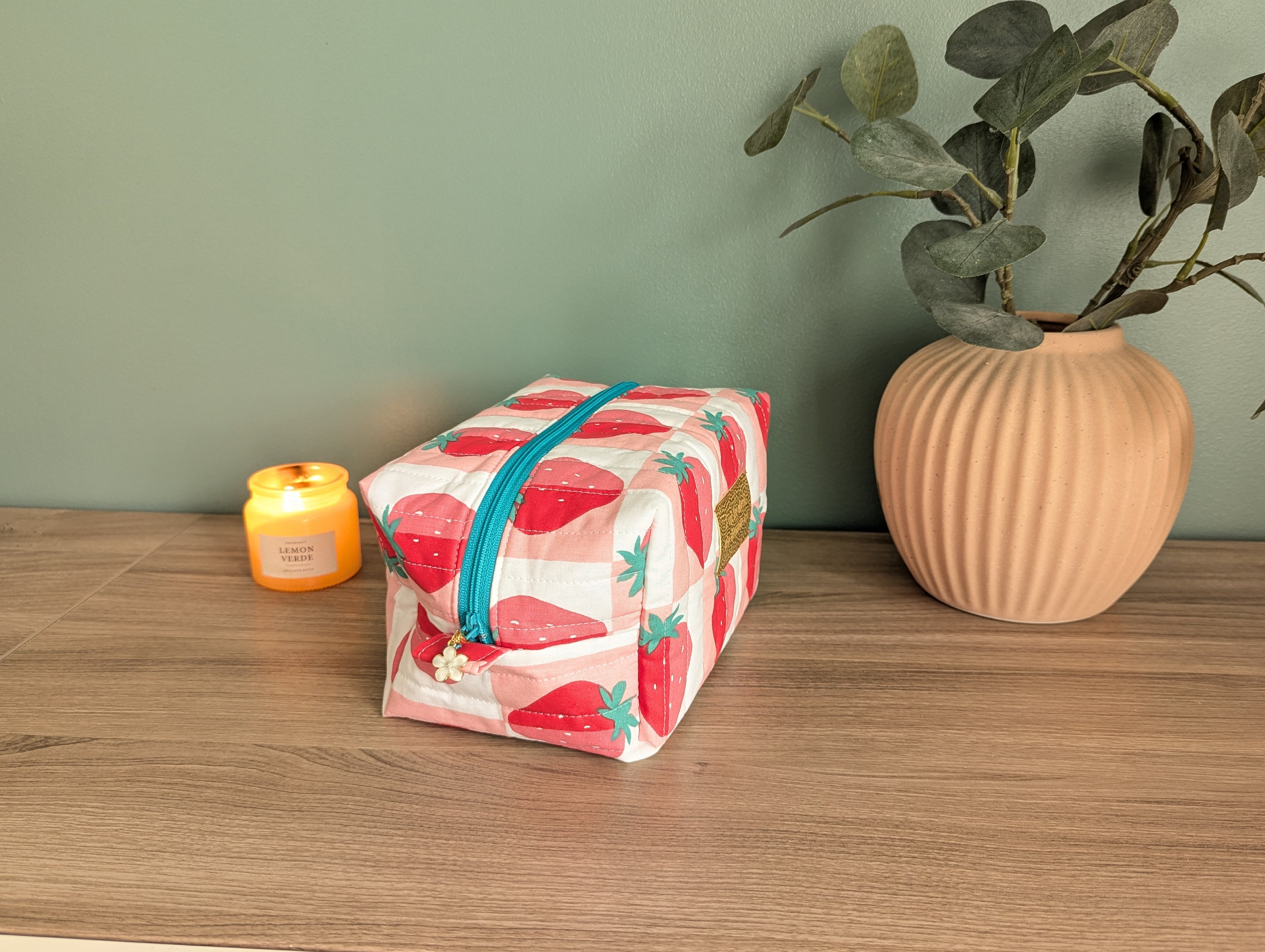 A pink and white striped cosmetic bag with a teal zipper sits on a wooden surface next to a small yellow candle and a beige vase with green leaves.