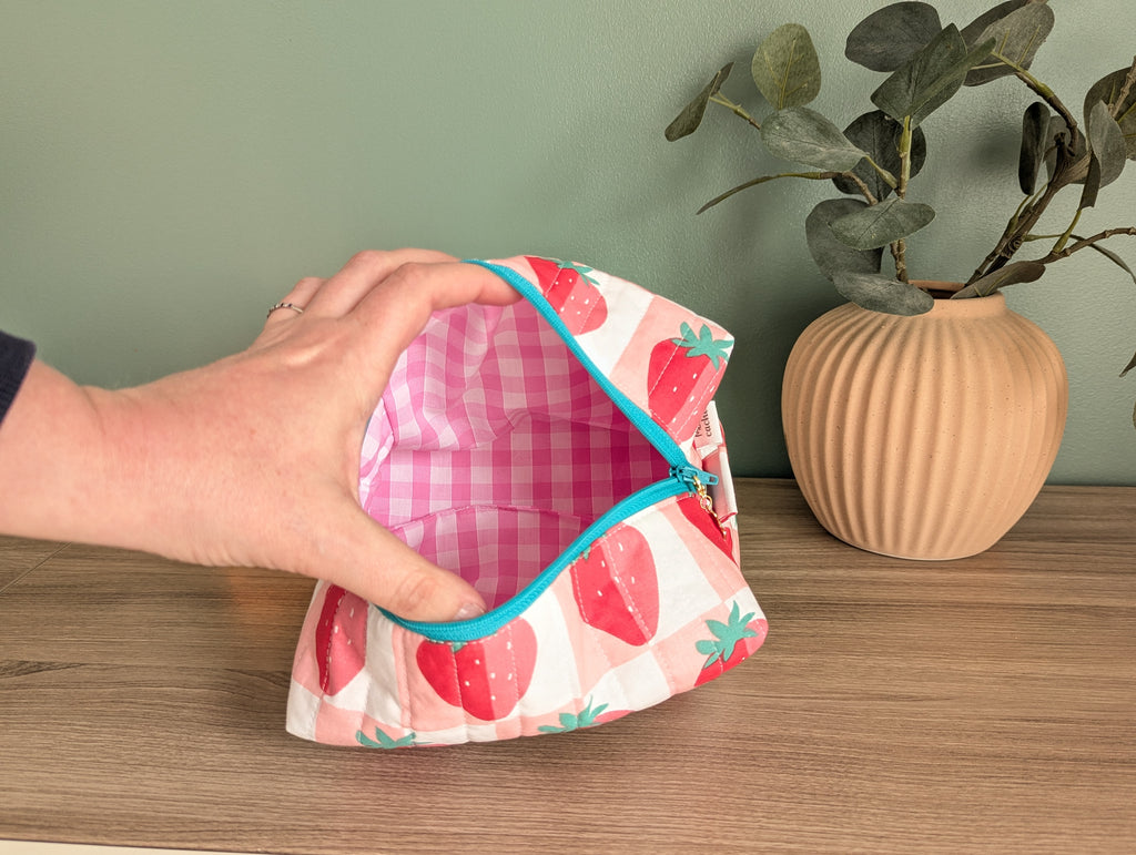 A hand is holding an open, colorful pouch with a strawberry pattern, placed on a wooden surface next to a potted plant.