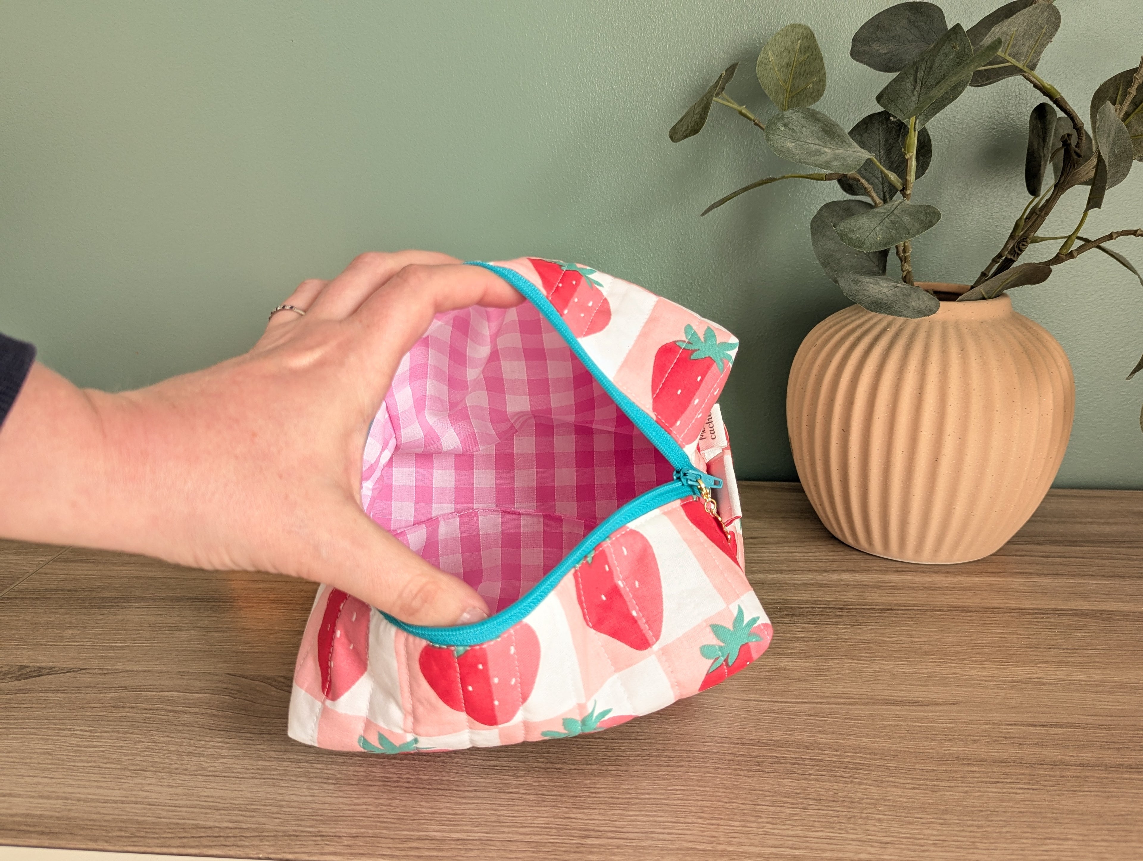 A hand is holding an open, colorful pouch with a strawberry pattern, placed on a wooden surface next to a potted plant.