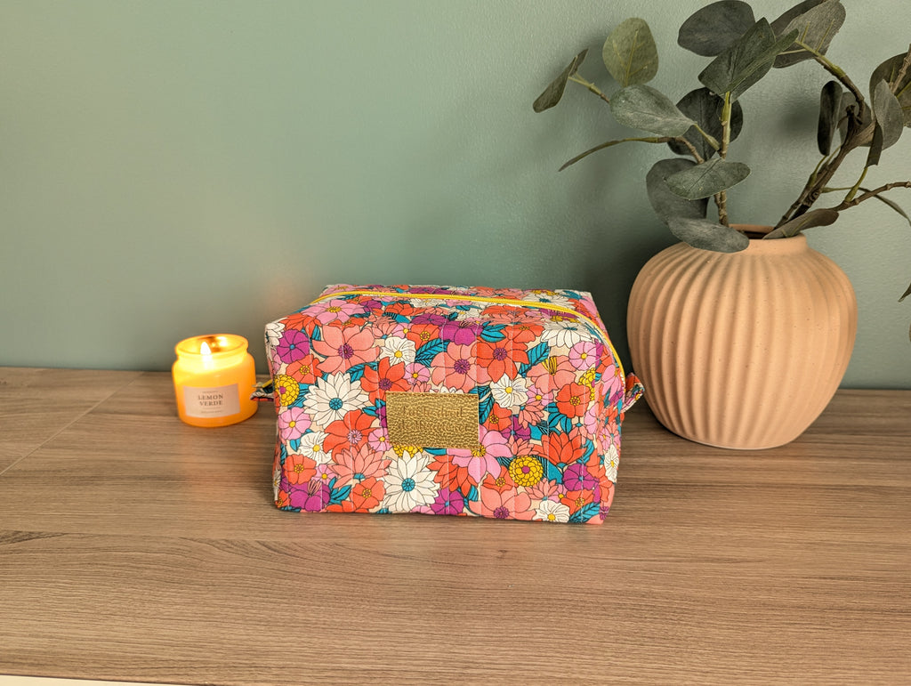 A colorful floral makeup bag sits on a wooden surface, accompanied by a small candle and a potted plant.